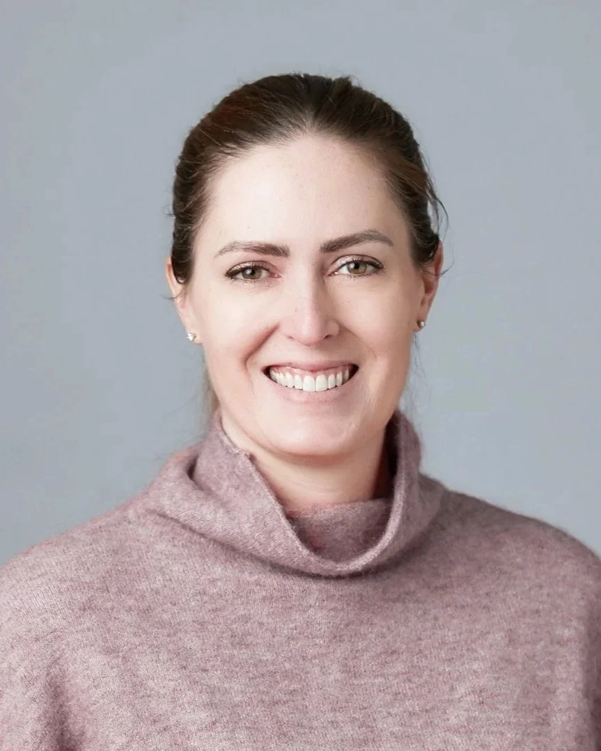 A woman with brown hair pulled back, wearing a pinkish-brown turtleneck sweater, smiling at the camera against a gray background.