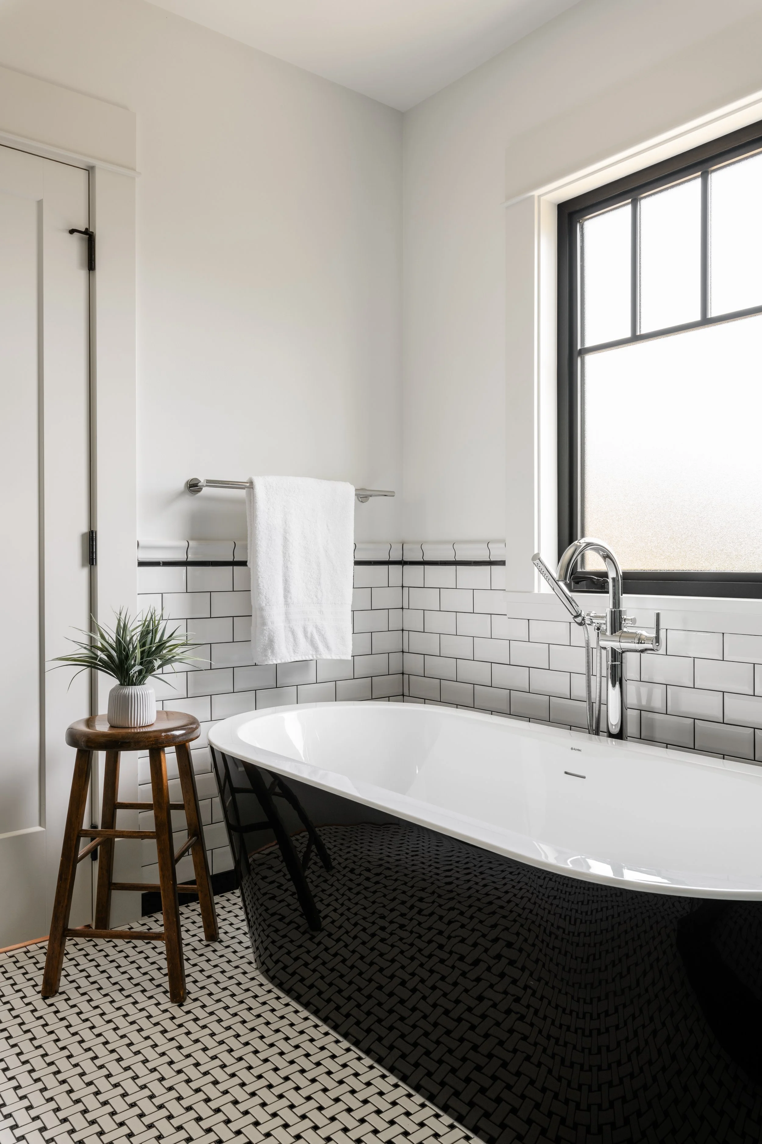 Modern bathroom with a black clawfoot bathtub next to a large frosted window, a small wooden stool with a potted plant, white subway tile wainscoting, and a white towel on a metal rack.