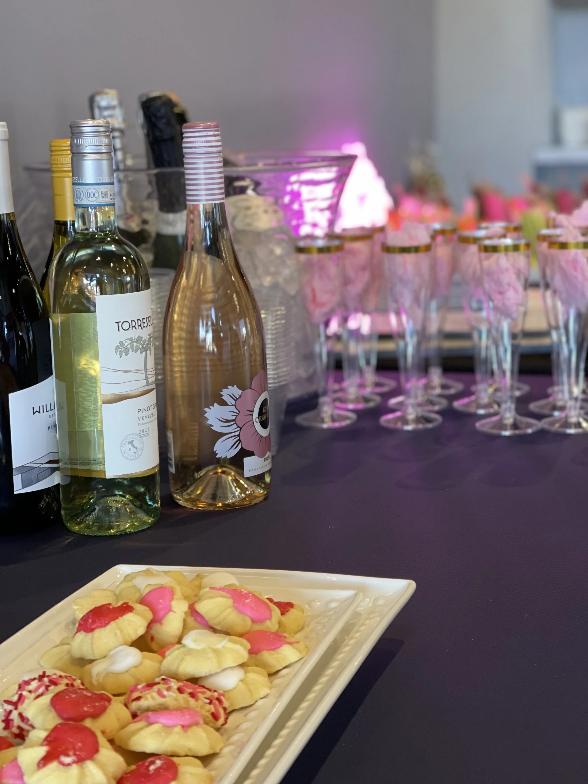 A table setup with wine bottles, glasses with pink decoration inside, and a tray of decorated cookies at a celebration event.