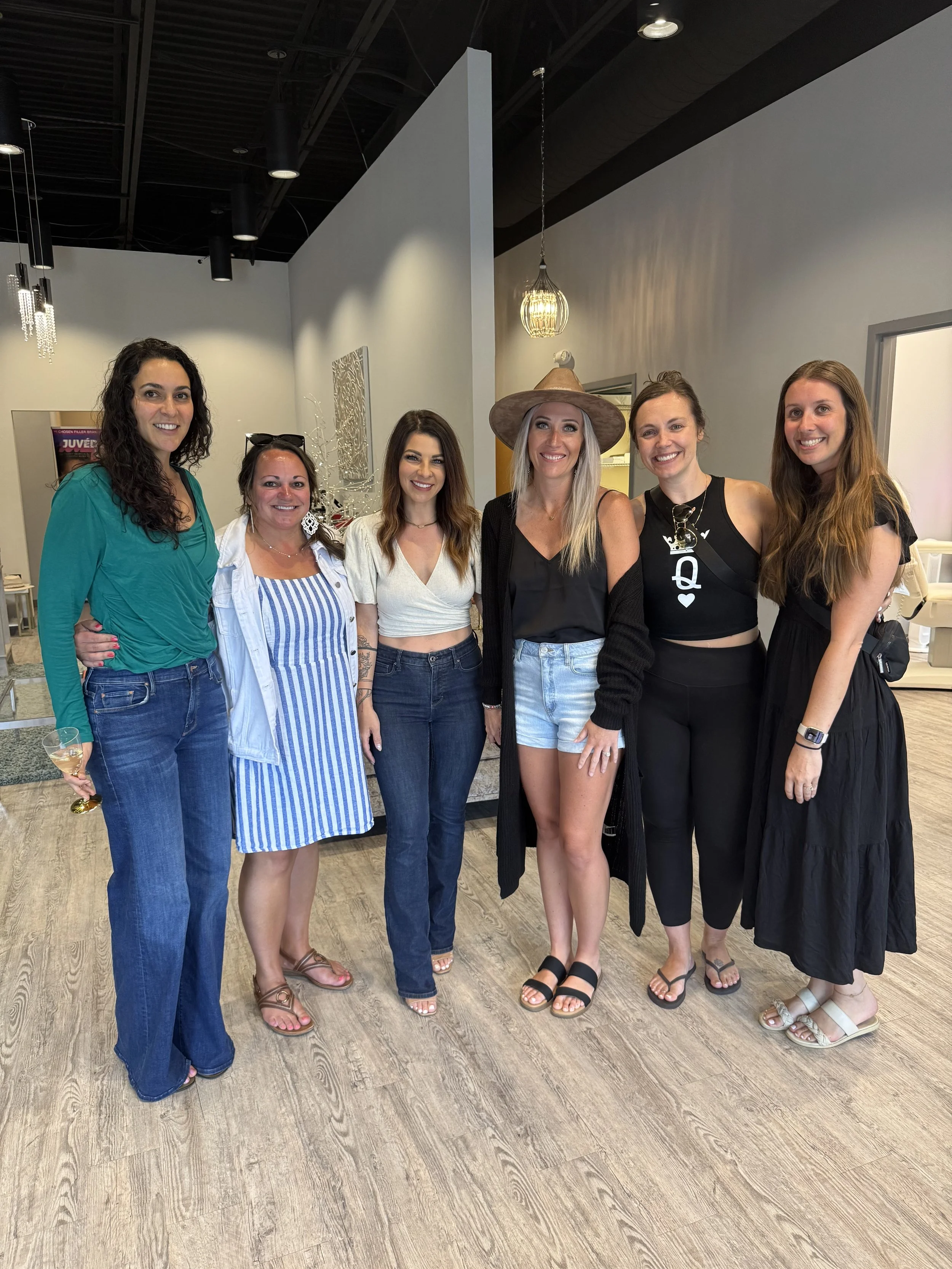 Six women standing together indoors, smiling at the camera. One woman is holding a glass of wine. They are dressed casually, with some wearing sandals and one wearing a wide-brimmed hat.