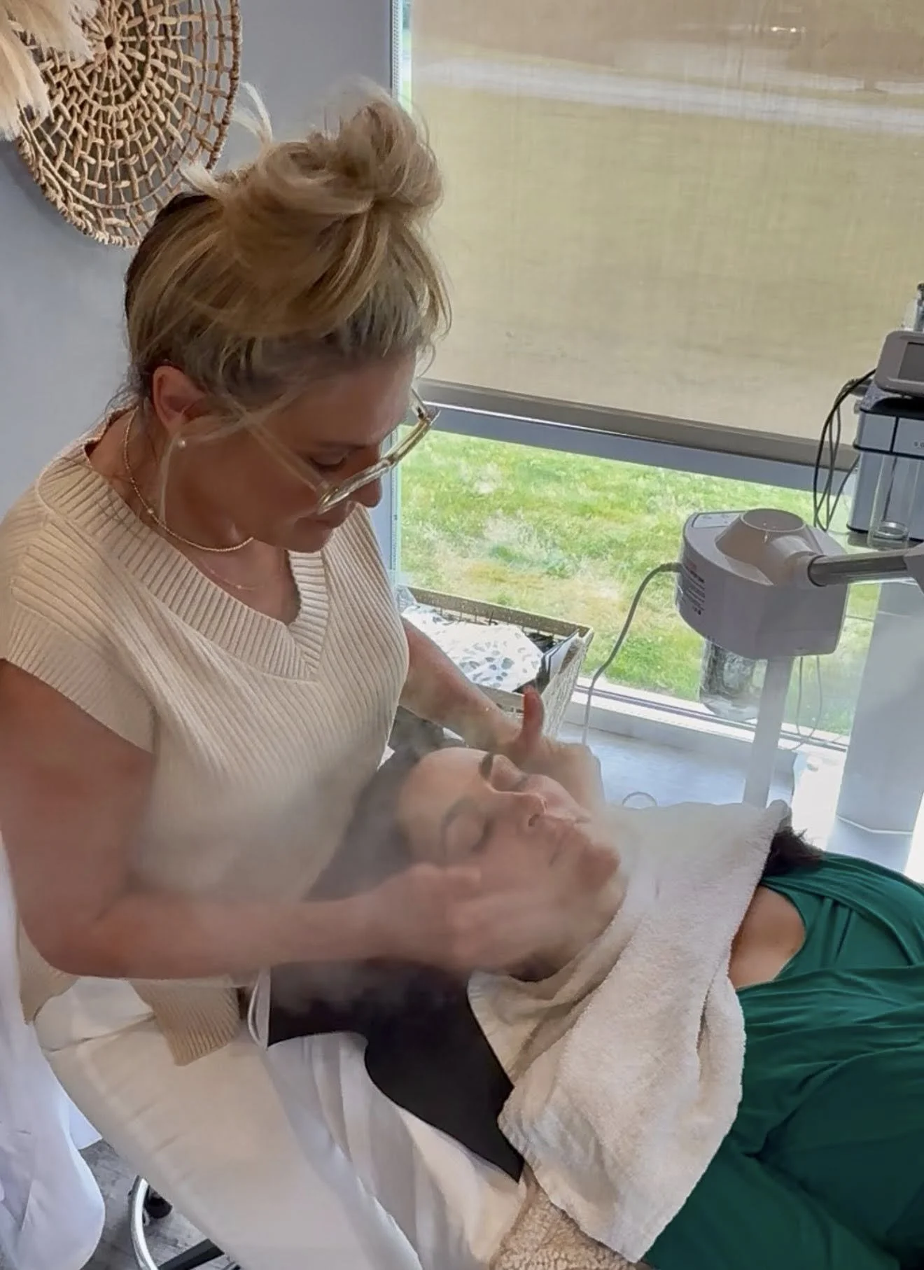 A woman wearing glasses and a beige top is giving a facial treatment to a woman lying on a treatment table with her eyes closed in a spa or skincare clinic.