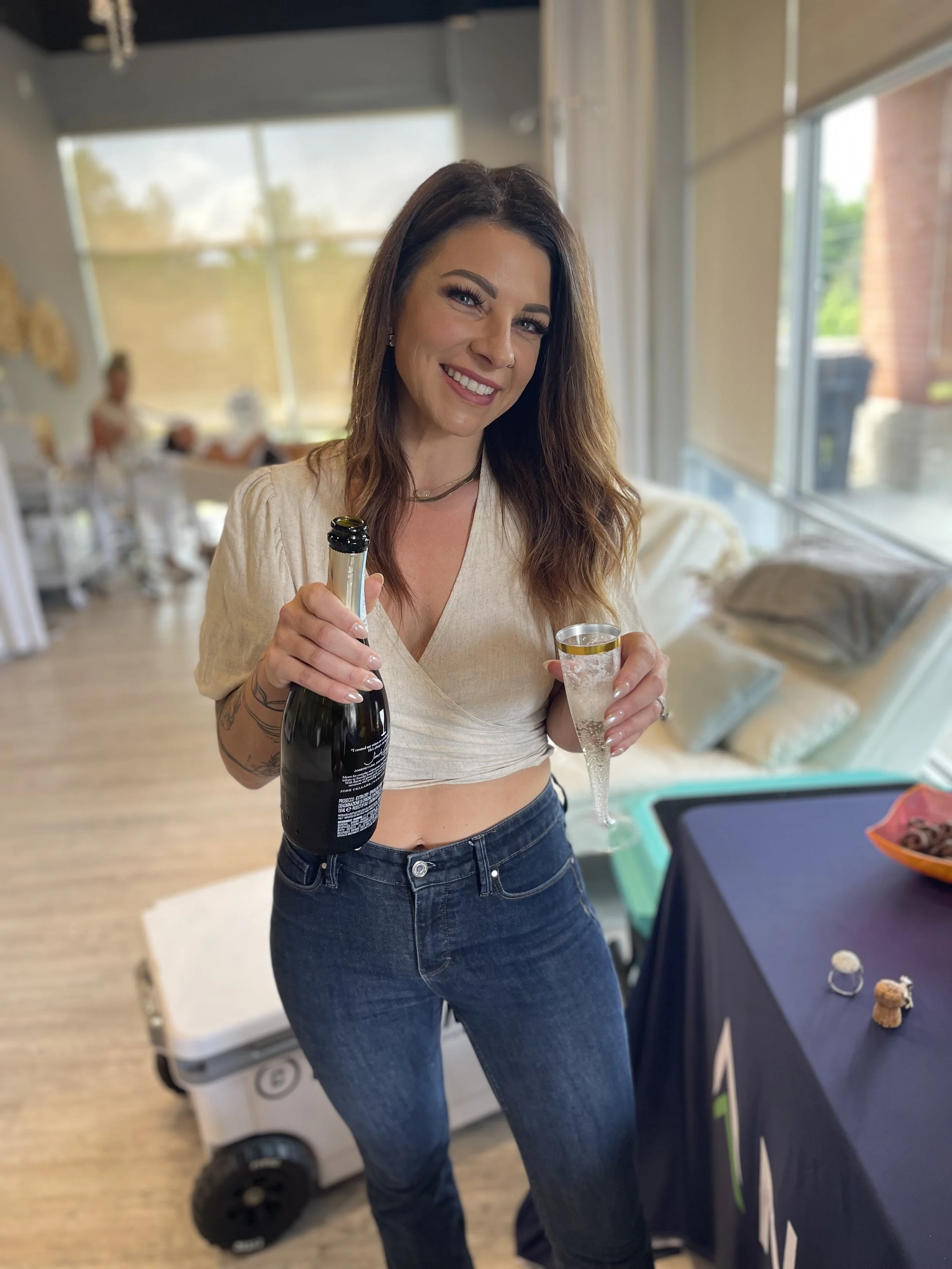 A young woman with long brown hair smiling and holding a bottle of champagne and a champagne flute in a bright, modern room. She is wearing a cream-colored crop top and dark jeans. In the background, there are other people and a large window with outdoor views.