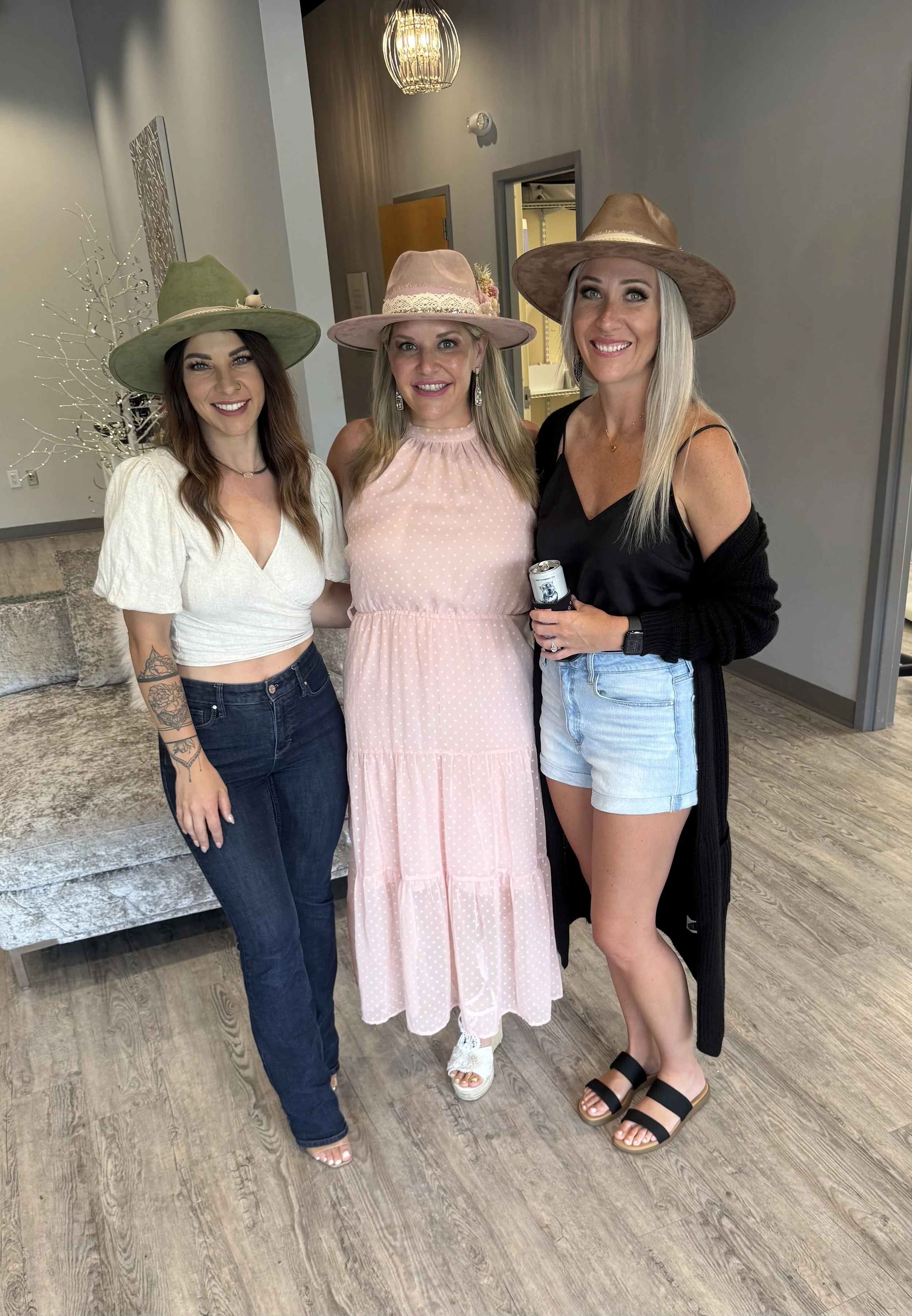 Three women standing together indoors, wearing large hats, with casual and summer clothing, smiling at the camera.