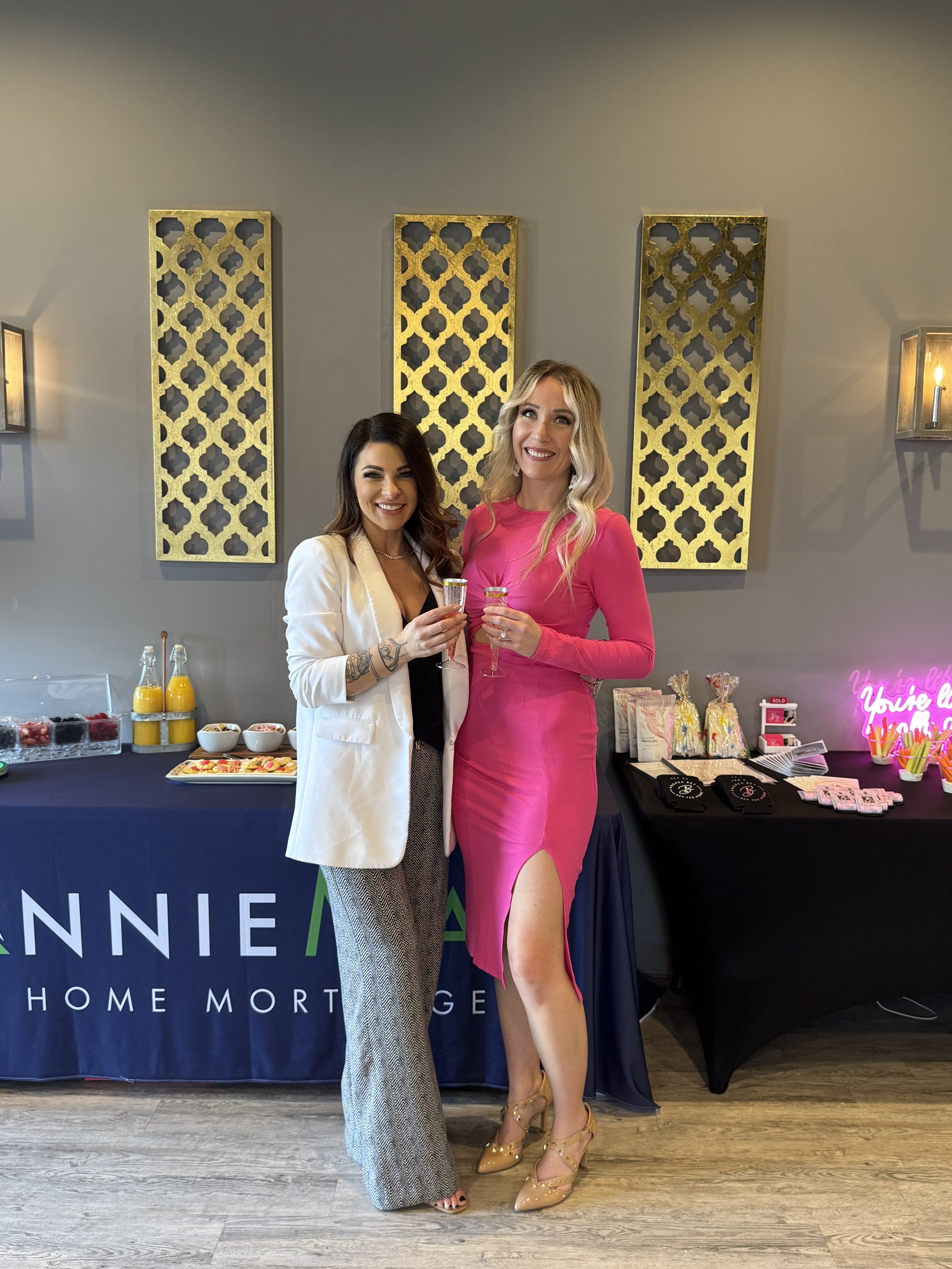 Two women standing together at a social event, smiling and holding drinks, in front of a table with snacks, and a backdrop with decorative gold panels behind them.