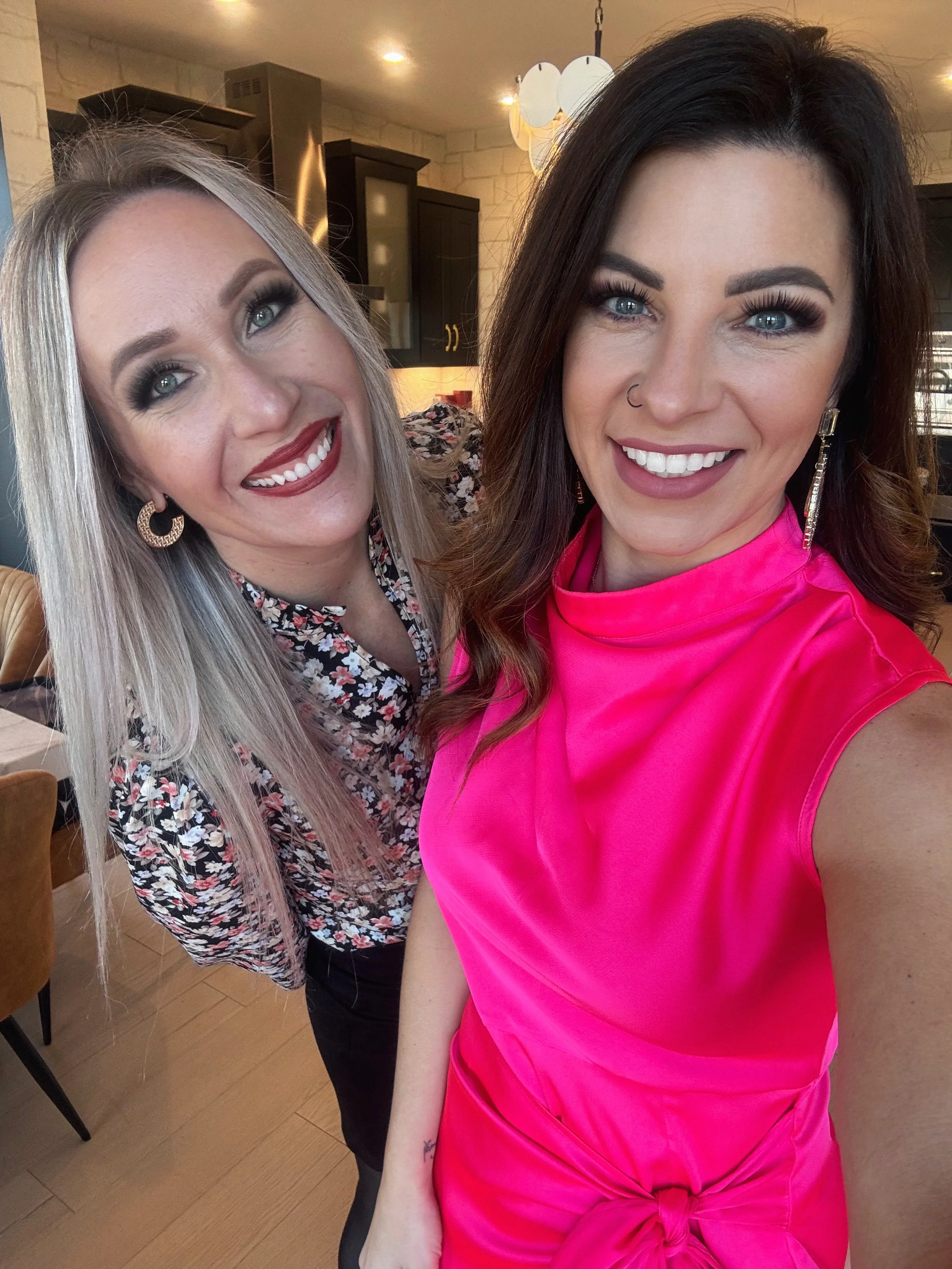 Two women smiling for a selfie in a kitchen area, one with blonde hair wearing a floral top and hoop earrings, the other with dark hair wearing a bright pink sleeveless top and long earrings.