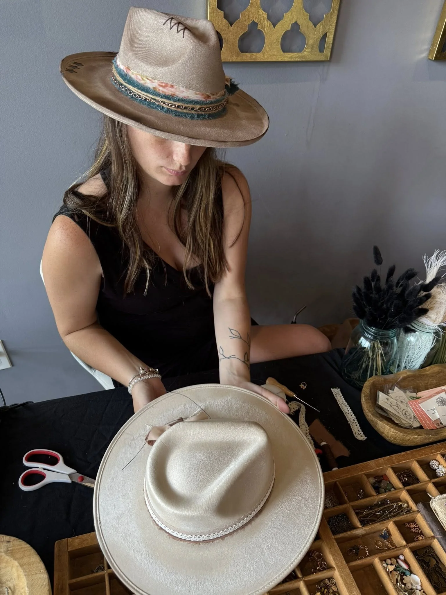 A woman sitting at a table wearing a large tan hat with colorful accents, surrounded by jewelry and craft supplies.