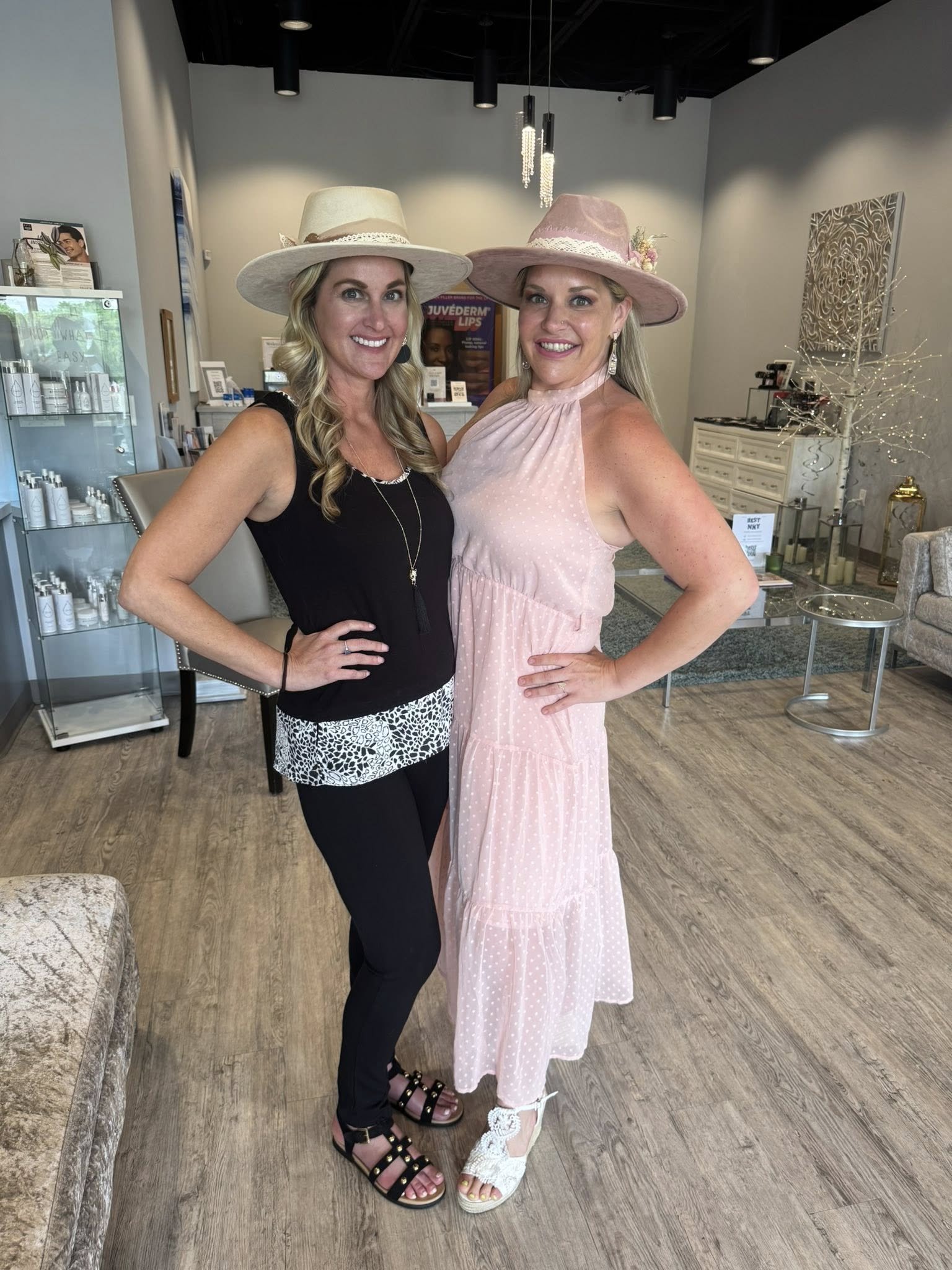 Two women standing inside a store, both wearing large hats and smiling at the camera. One woman is dressed in a pink dress, and the other in a black top and patterned white and black pants.