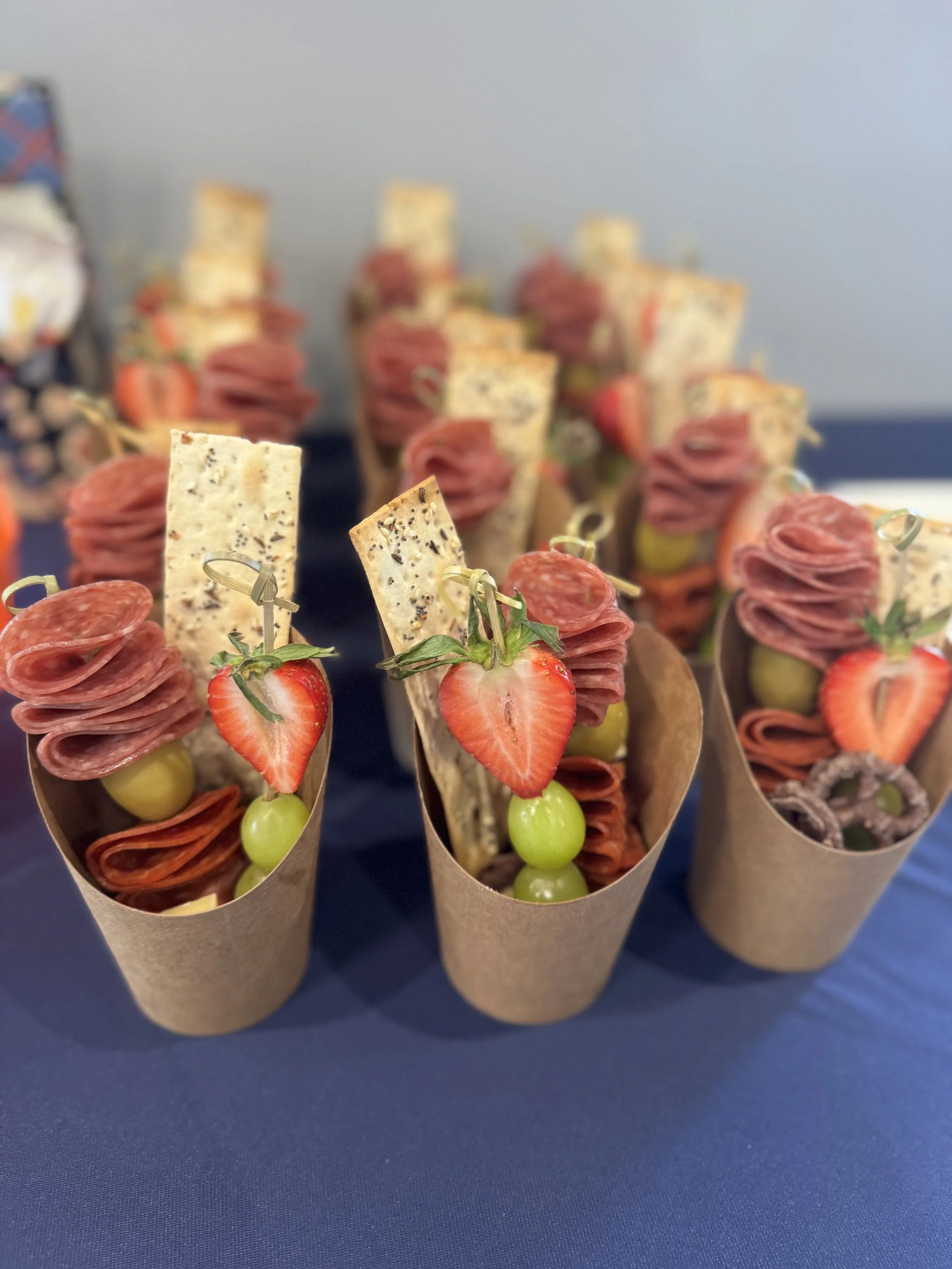 Appetizer servings with slices of cured meat, strawberries, green grapes, and cheese, arranged in small paper cones on a blue table.
