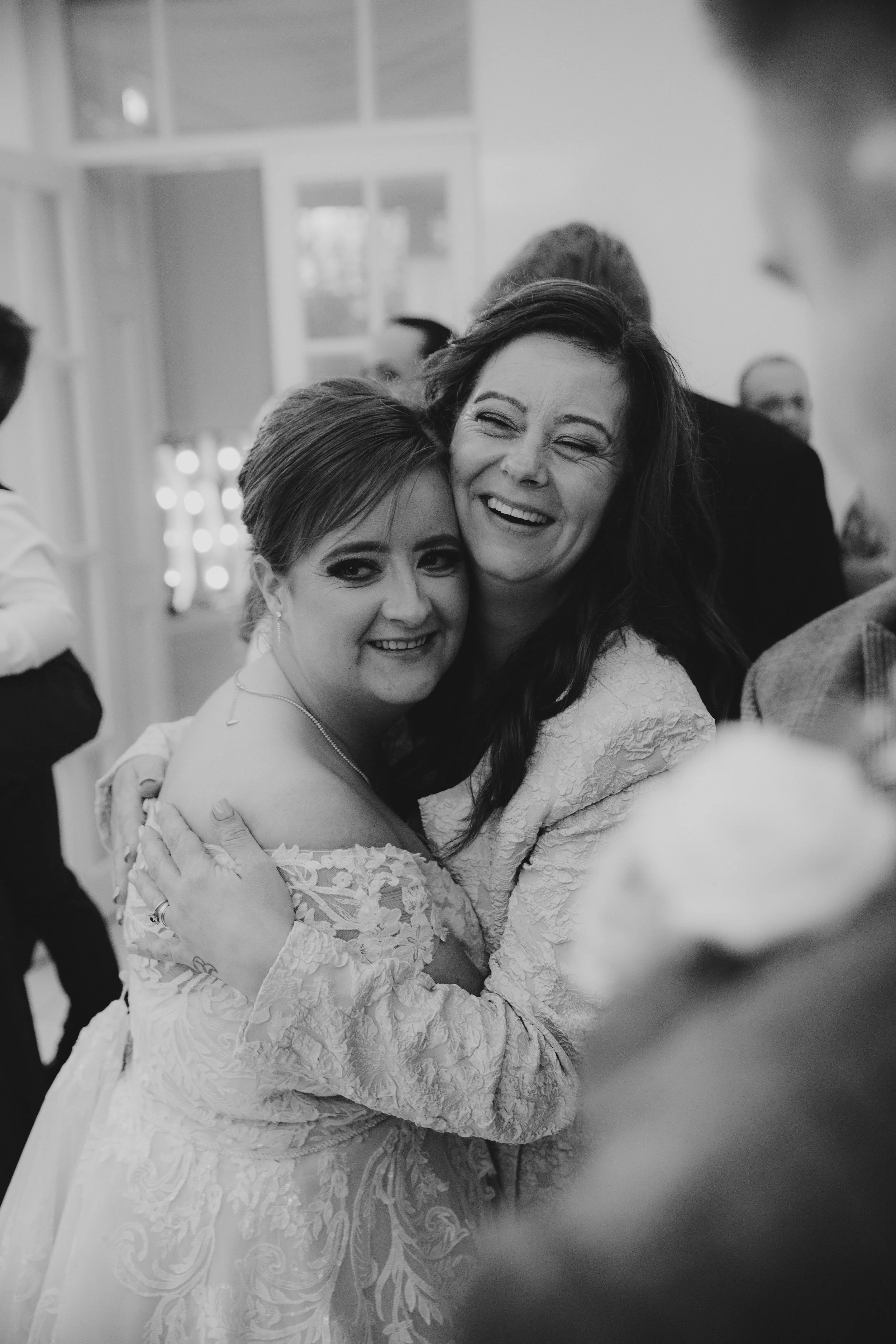 A black and white photograph of two women hugging and smiling, with a blurred background of people and a decorated wall, at a social event or celebration.