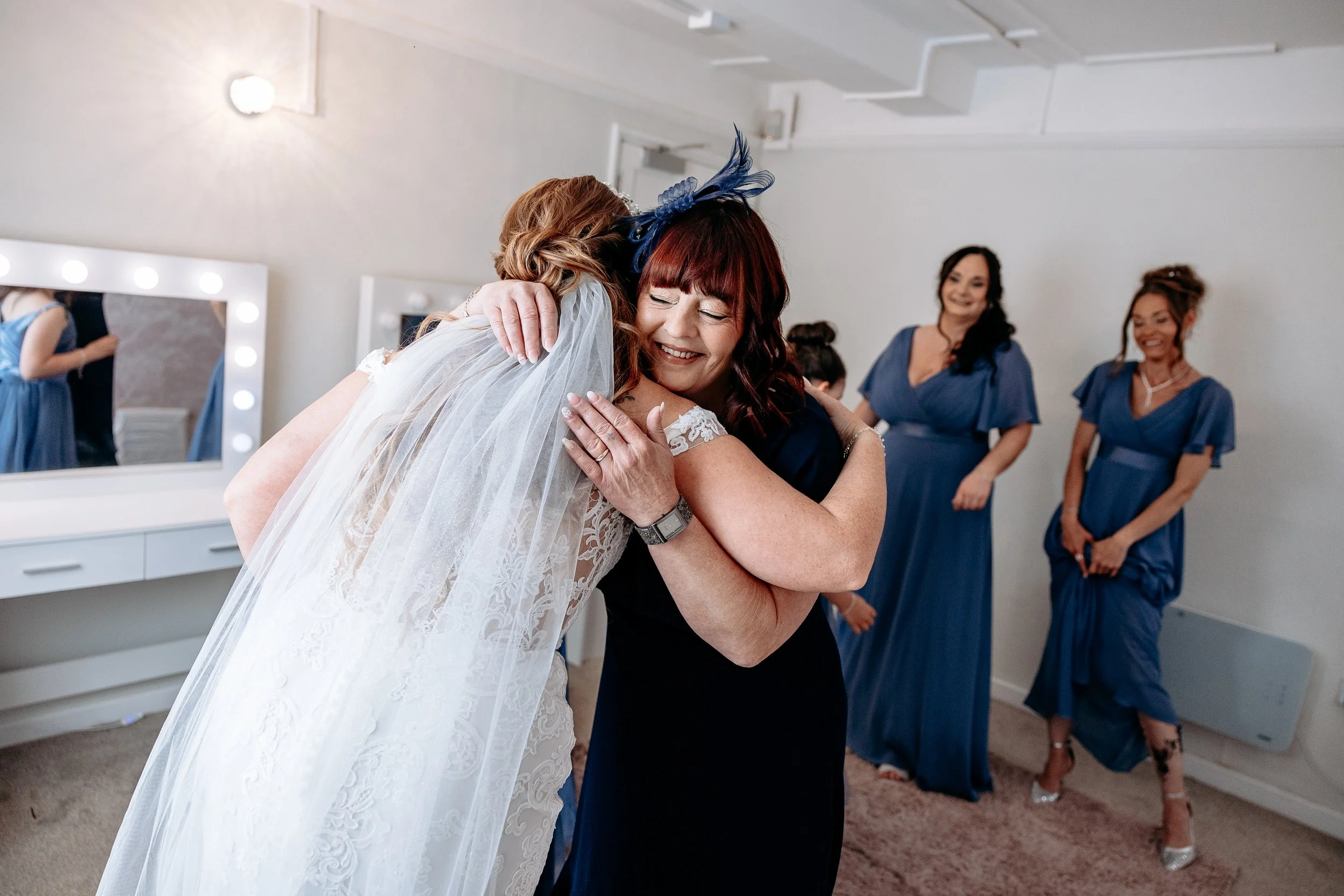 A bride and an older woman hug each other happily in a bridal suite, with three women in matching blue dresses smiling in the background.