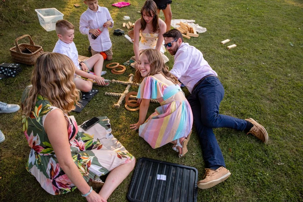 A group of children and adults playing Jenga and outdoor games on grass at a picnic or gathering.