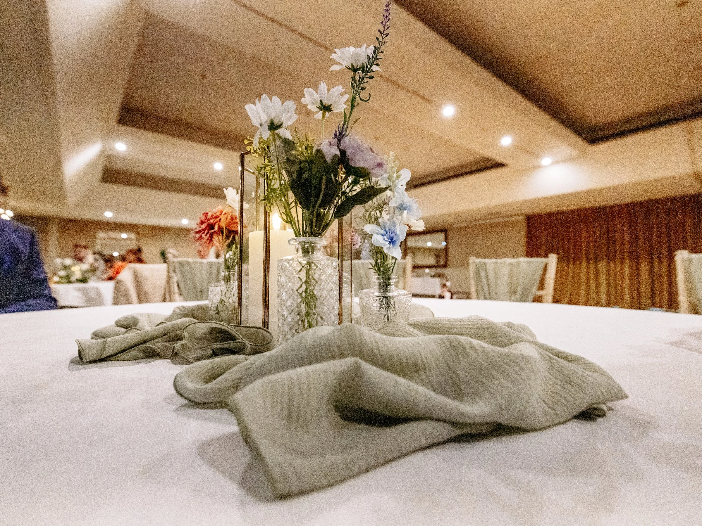 A centerpiece with three glass vases filled with white and light blue flowers on a dining table, with wrapped napkins and candles inside the vases, in an elegant banquet hall.