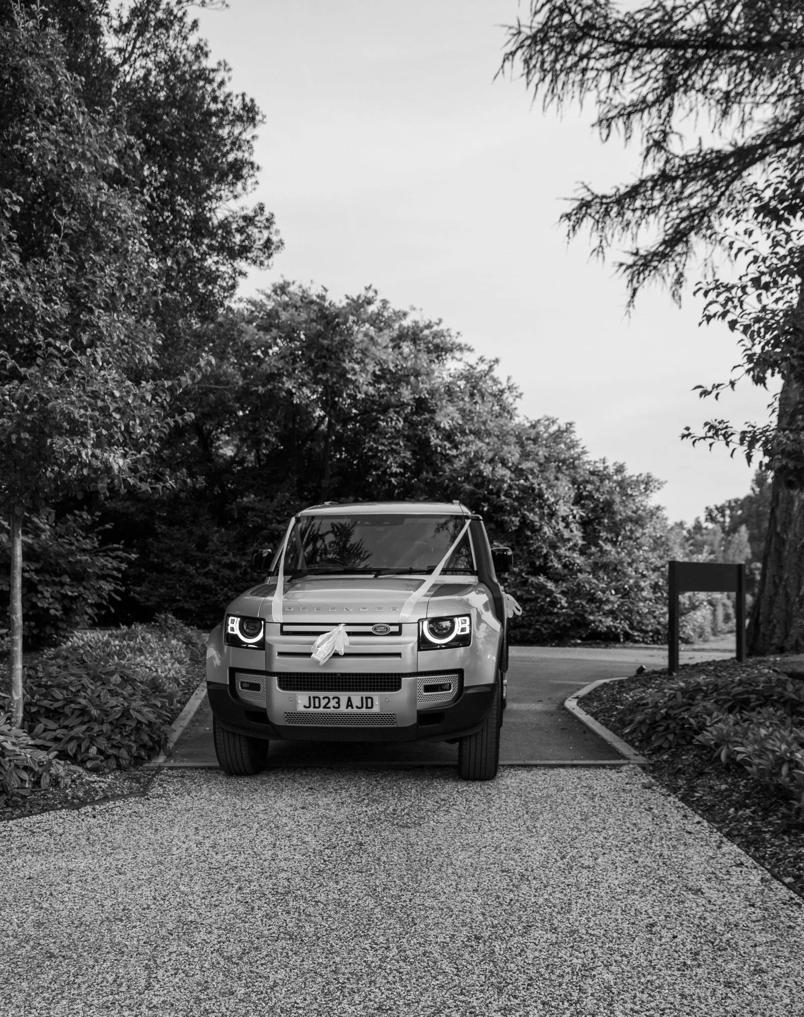 Black and white image of a Land Rover Defender decorated with ribbons, parked on a gravel path, surrounded by trees and greenery.
