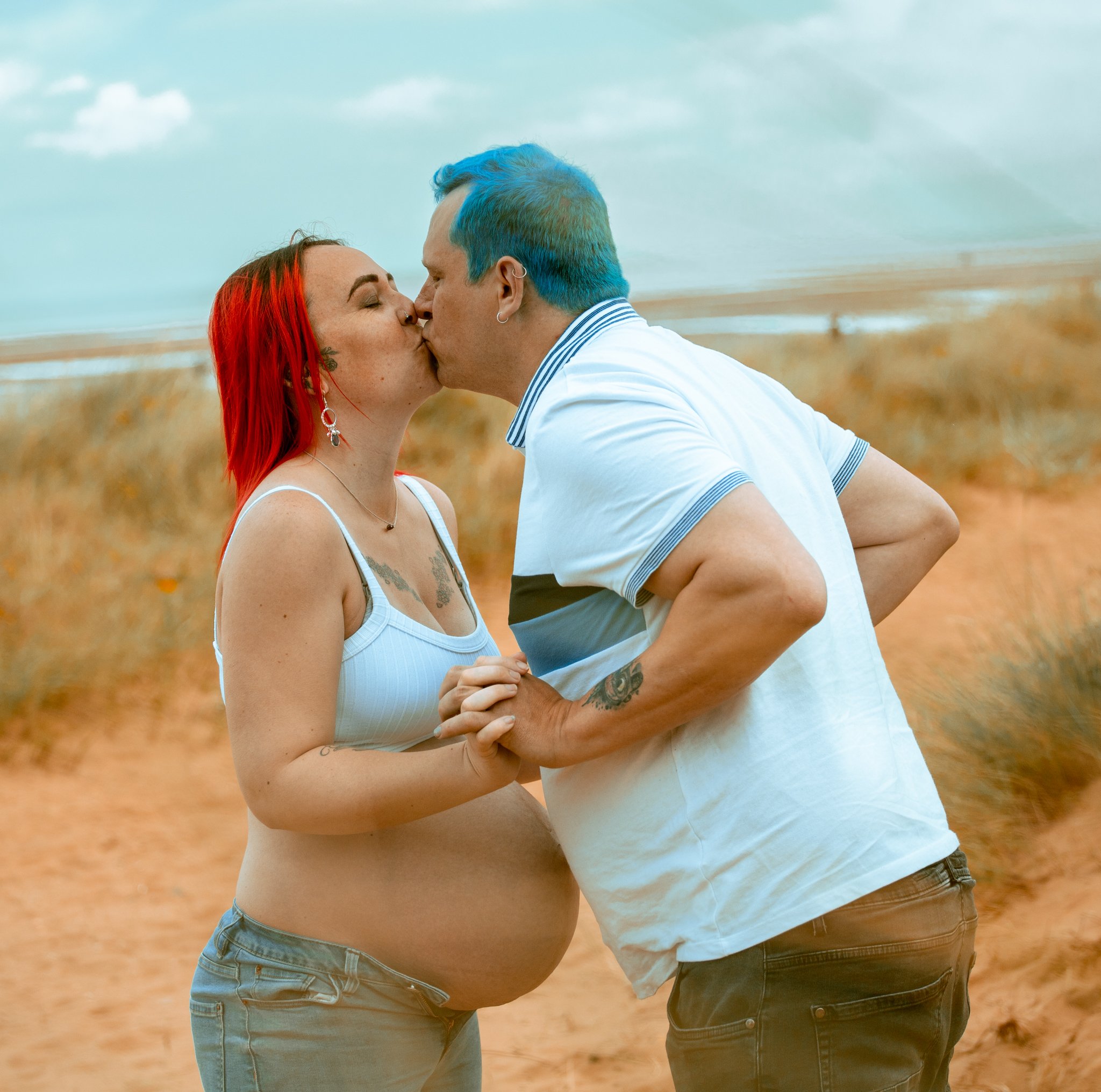 A pregnant woman with red hair and a tattoo on her chest, and a man with colorful blue hair, are holding hands and kissing on a beach with a cloudy sky in the background.