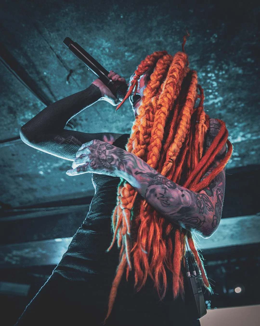 A person with orange dreadlocks and tattooed arms performs on stage, holding a microphone and singing with a dark, textured background.
