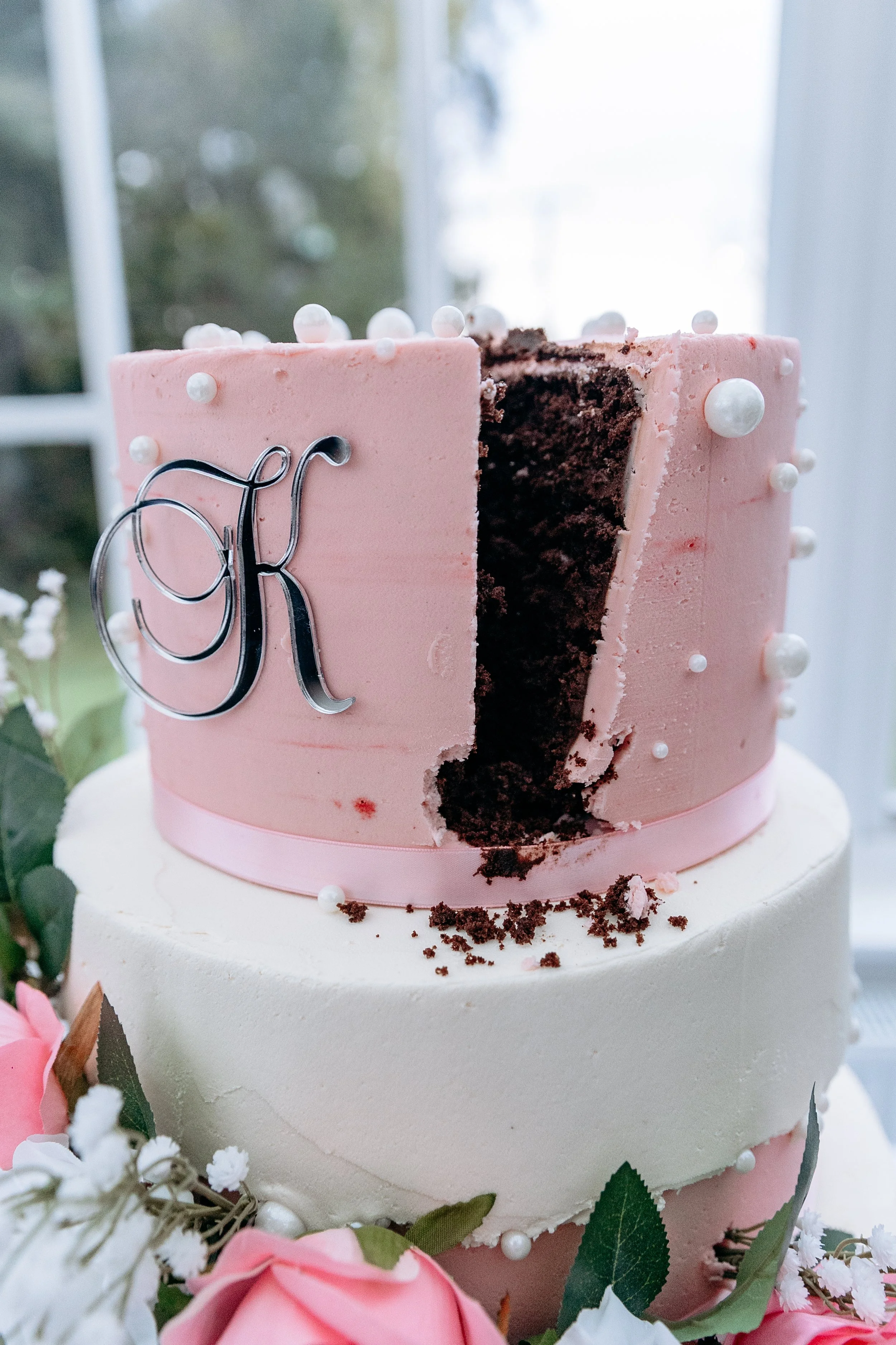 Two-tiered wedding cake with pink and white frosting, decorated with edible pearls, a metallic monogram letter 'K', and surrounded by pink roses and white flowers.