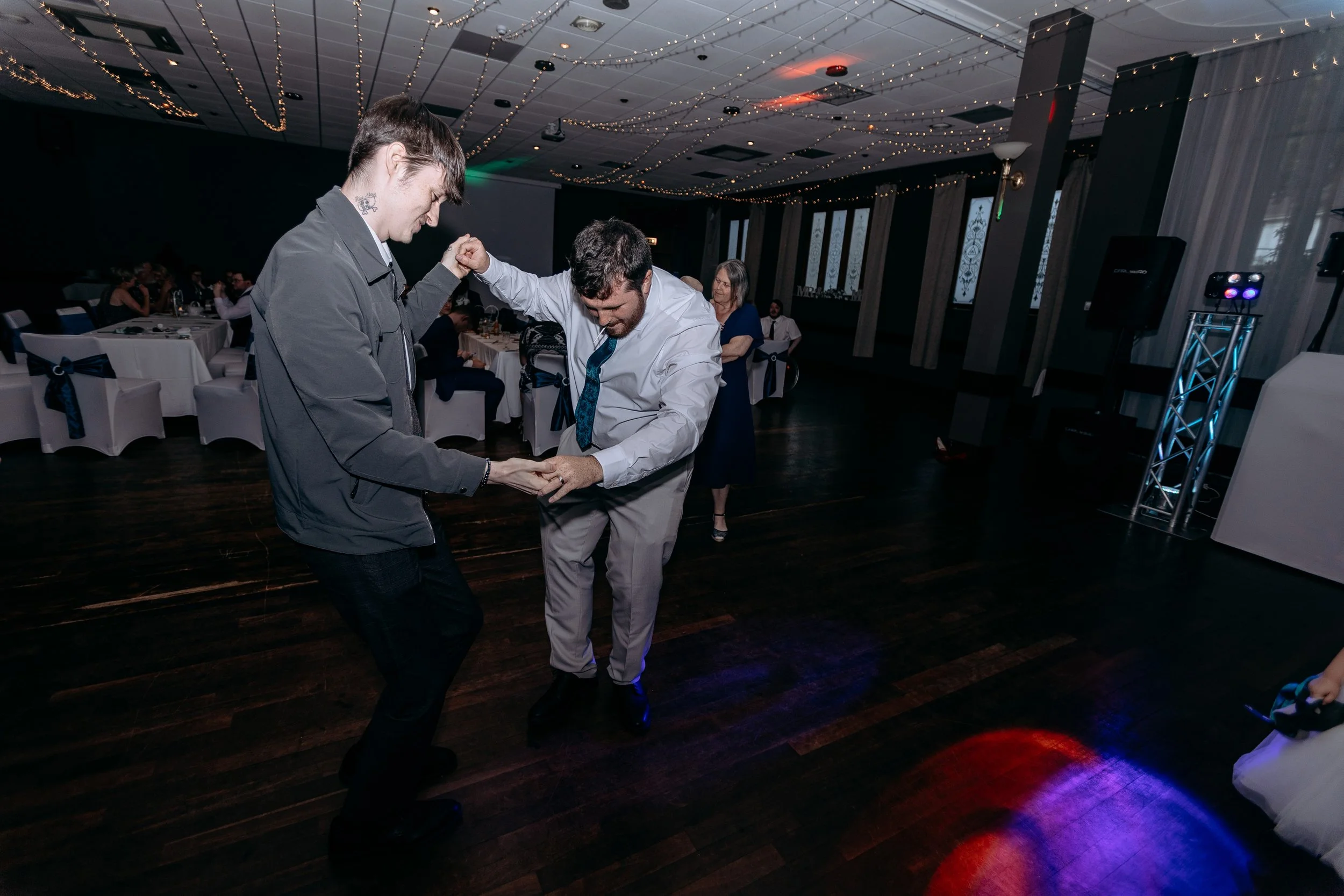 Two men are dancing and holding hands at a celebration or party with tables, chairs, and string lights in the background.