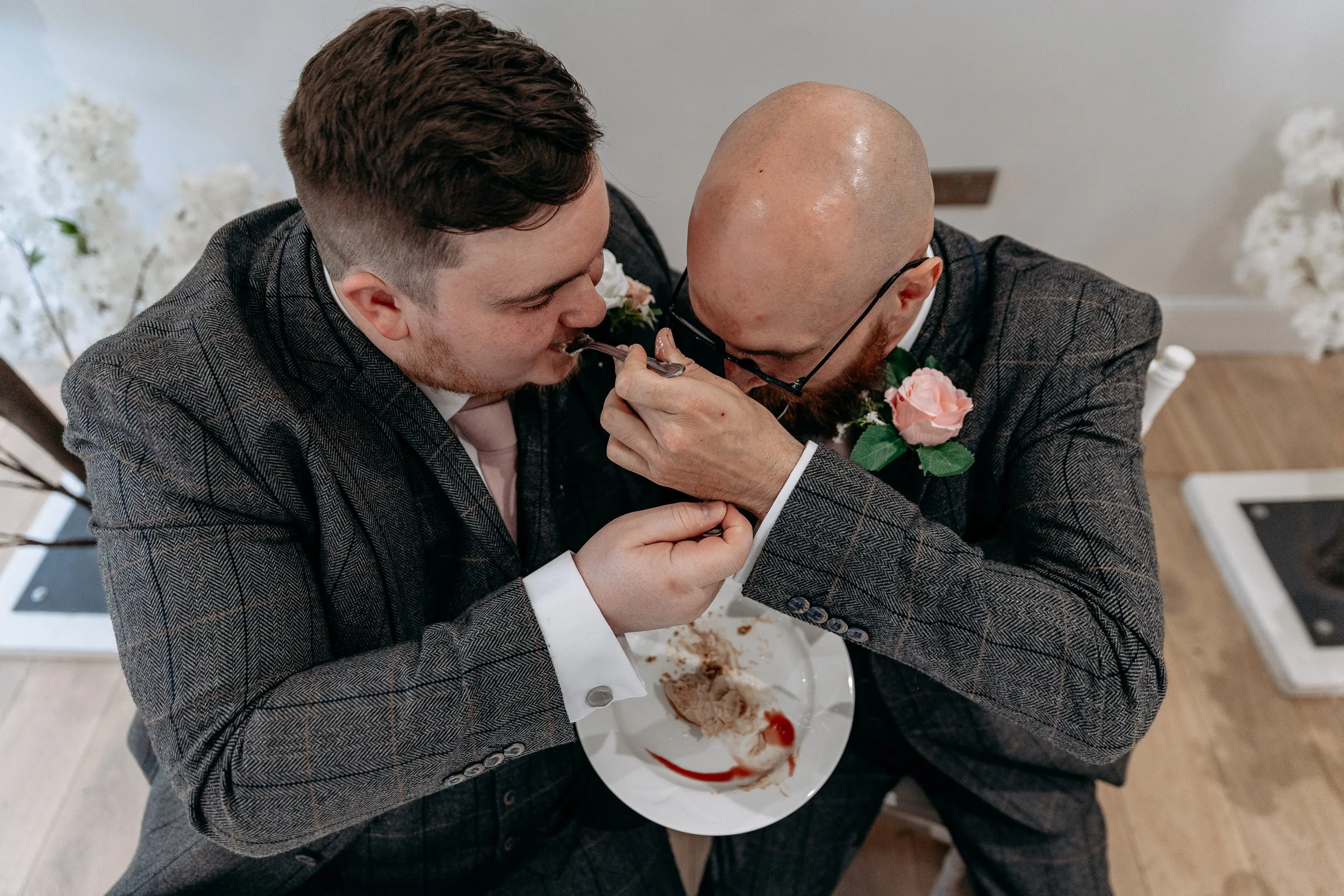 Two men in suits sharing a fork of cake at a wedding reception, one of them with a pink rose boutonniere.
