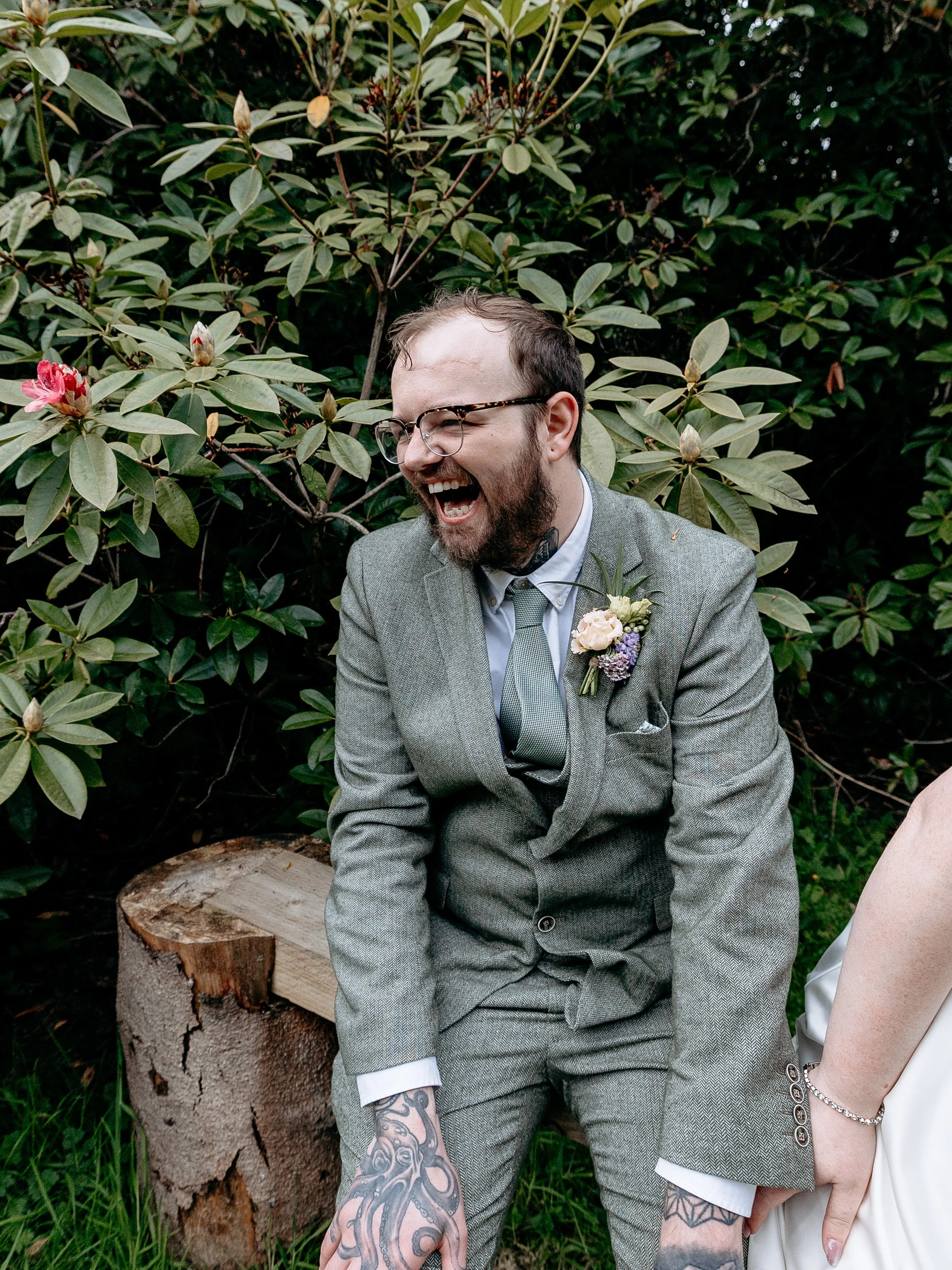 Man in gray suit laughing, sitting on a wooden bench, outdoors, surrounded by green bushes and flowering plants.