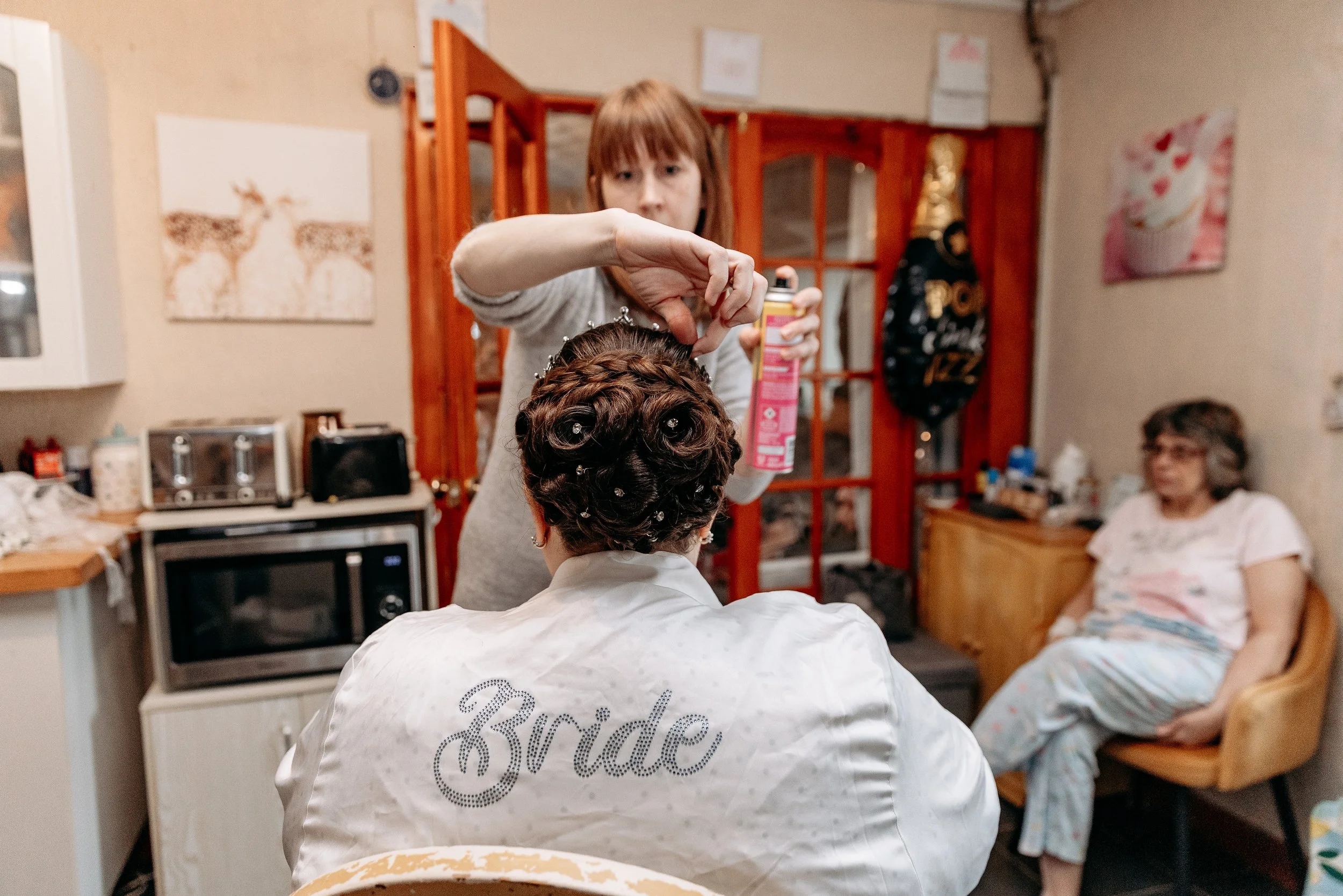 A bride having her hair styled with curls and hairpins in a cozy room, while a woman sits relaxed in a chair in the background.