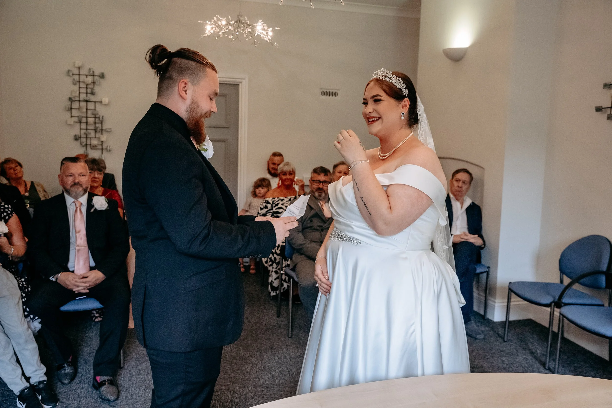 Bride and groom exchanging vows during wedding ceremony in a room with seated guests.