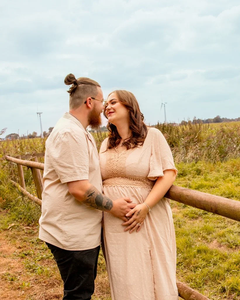 A couple standing close together outdoors, holding hands, with a scenic rural background including wind turbines and cloudy sky.
