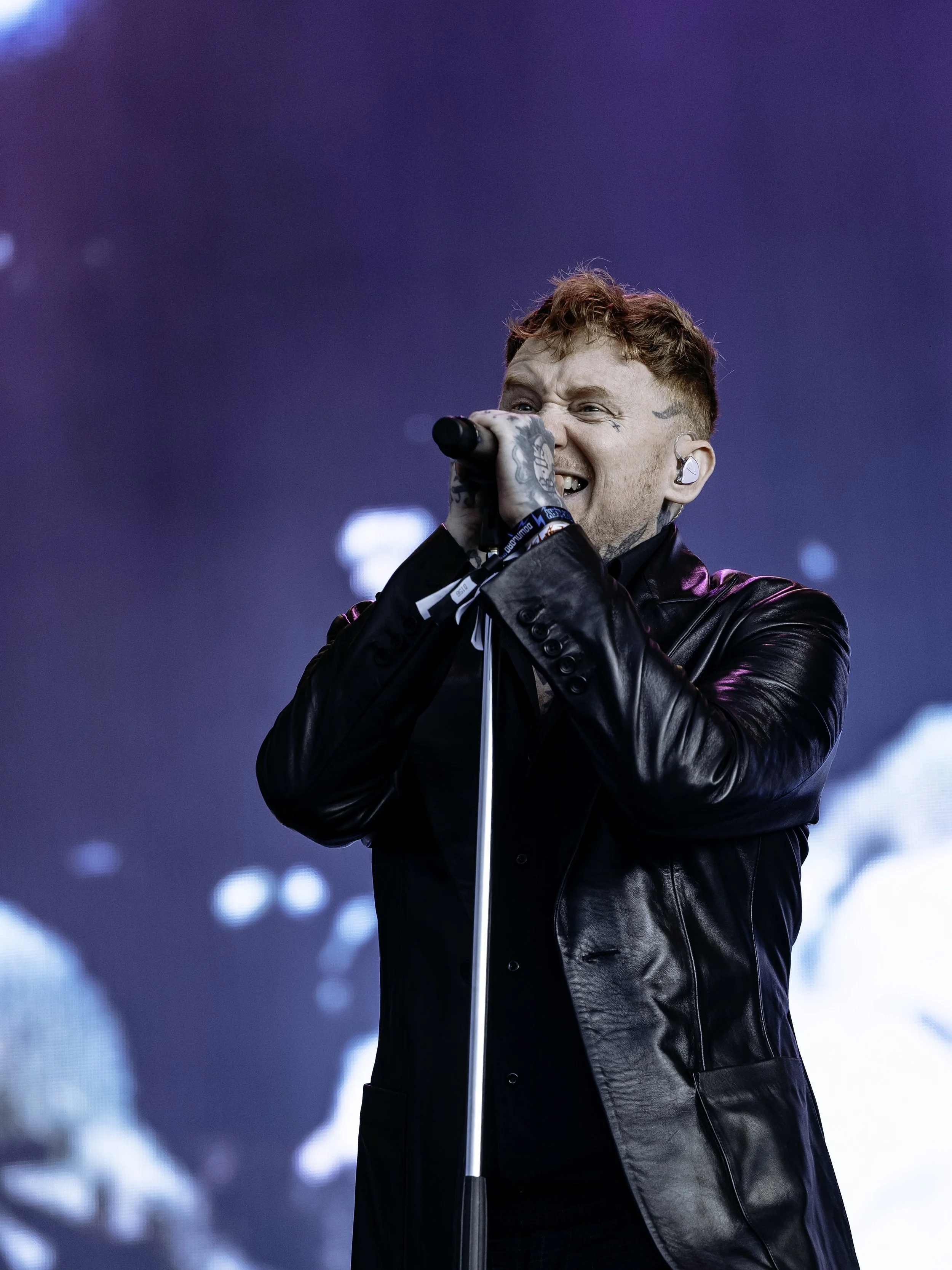 A male singer with red hair, tattoos, and wearing a black leather jacket, passionately singing into a microphone on stage.