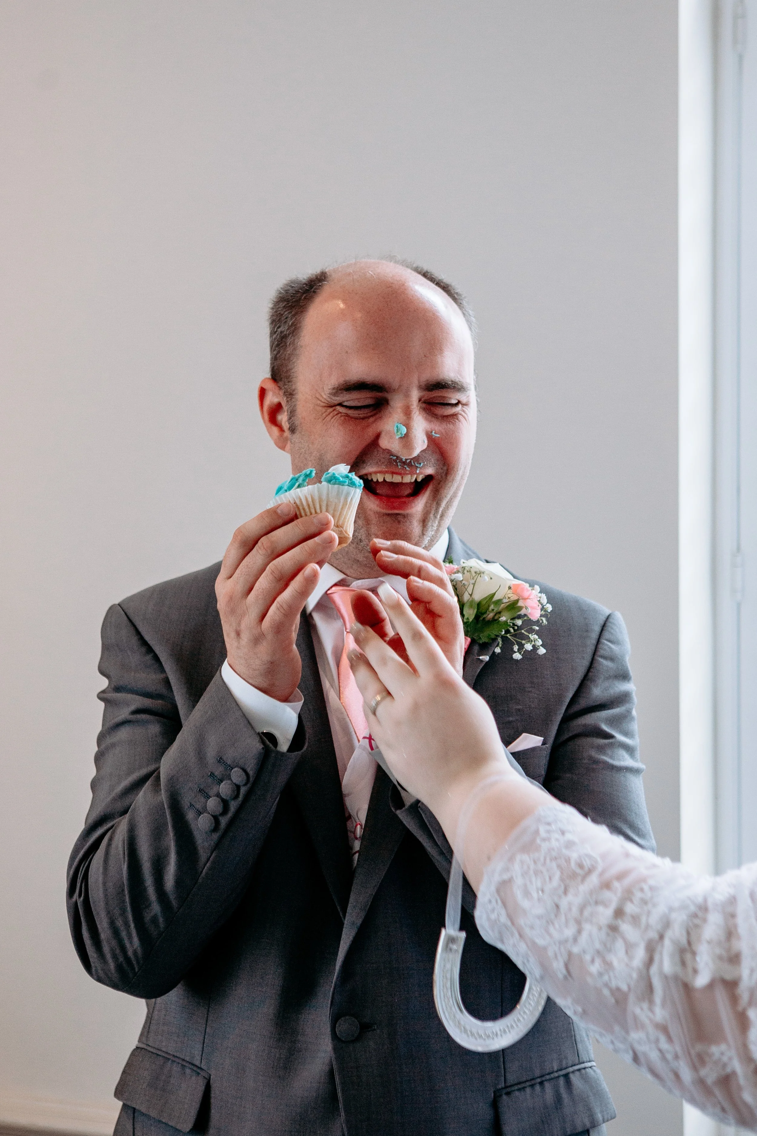 A man in a tuxedo with a boutonniere is smiling as he is fed a cupcake by a person wearing a white lace sleeve, with blue frosting on his nose and face.