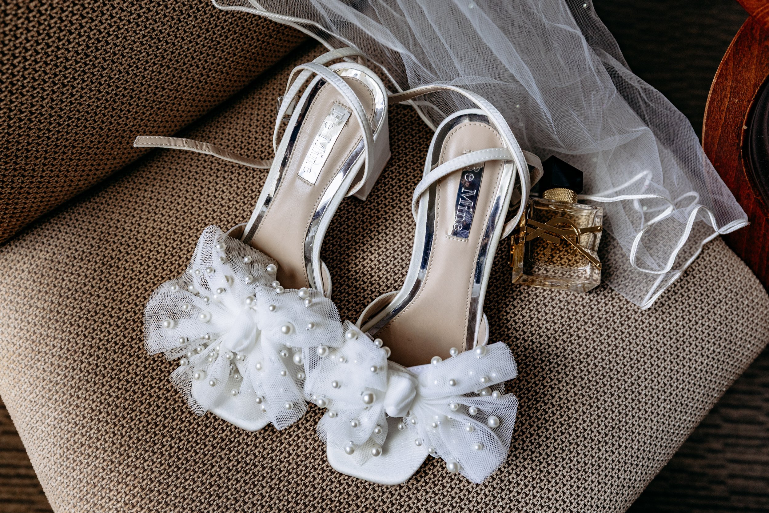 A pair of white high-heeled shoes with pearl-embellished bow decorations, placed on a brown textured cushion, with a bottle of perfume and a sheer veil nearby.
