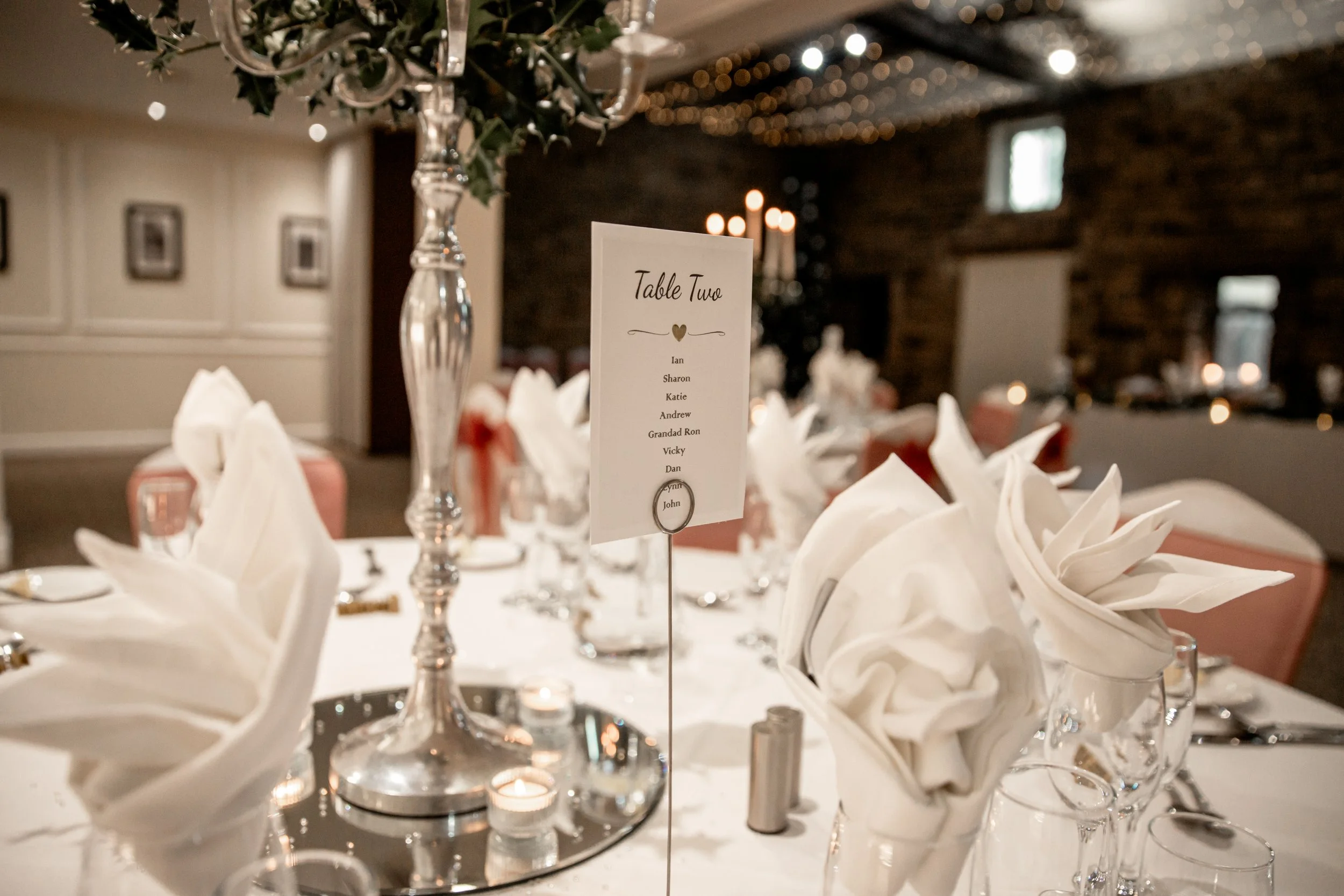 Elegant wedding reception table with white napkins folded in a fan shape, silverware, wine glasses, a tall silver candelabra, a table number card reading "Table Two" with a list of names, small candles, and pink chairs in a warmly lit room with brick