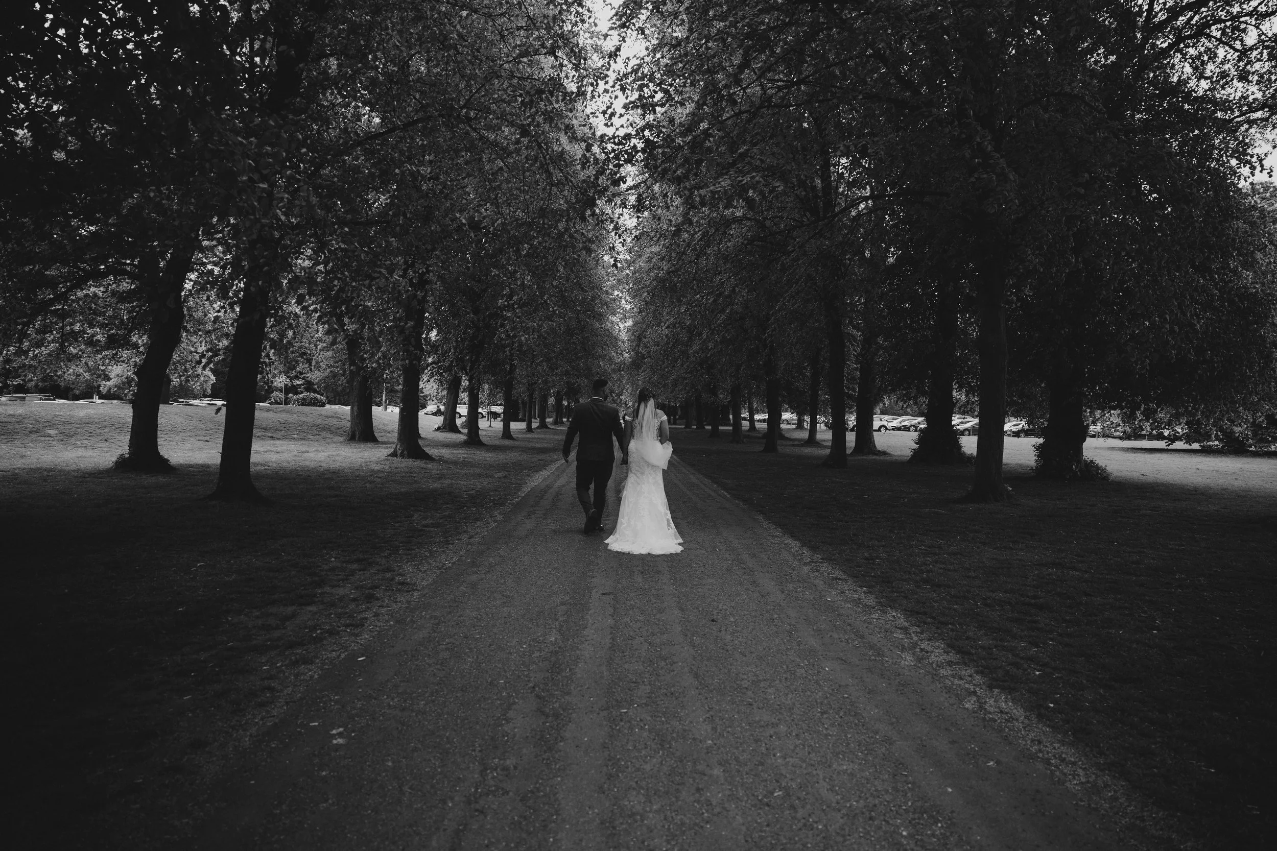 Black and white photo of a bride and groom walking down a tree-lined dirt path, holding hands, with parked cars visible in the background.