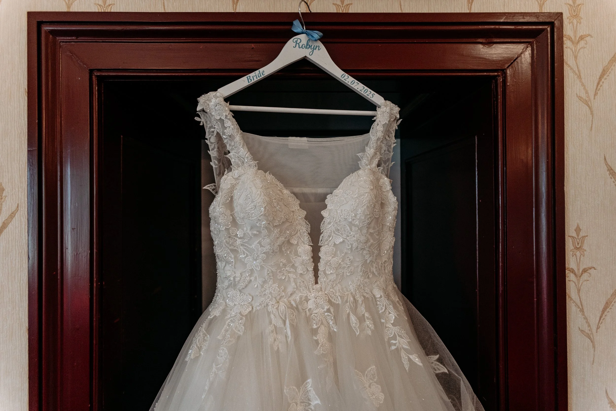 Wedding dress hanging on a white hanger in a wooden display, marked with the name 'Robyn', 'Bride', and the date '02.07.2025'.