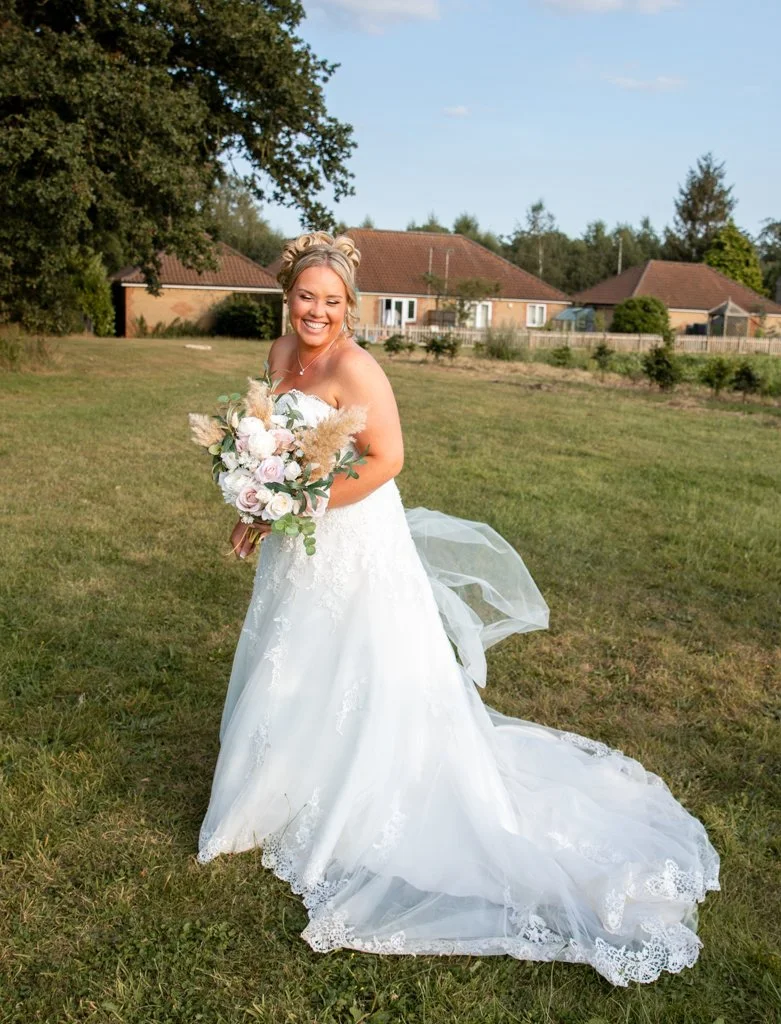 A smiling bride in a white wedding gown holding a bouquet of flowers, standing on a grassy lawn with houses and trees in the background under a partly cloudy sky.