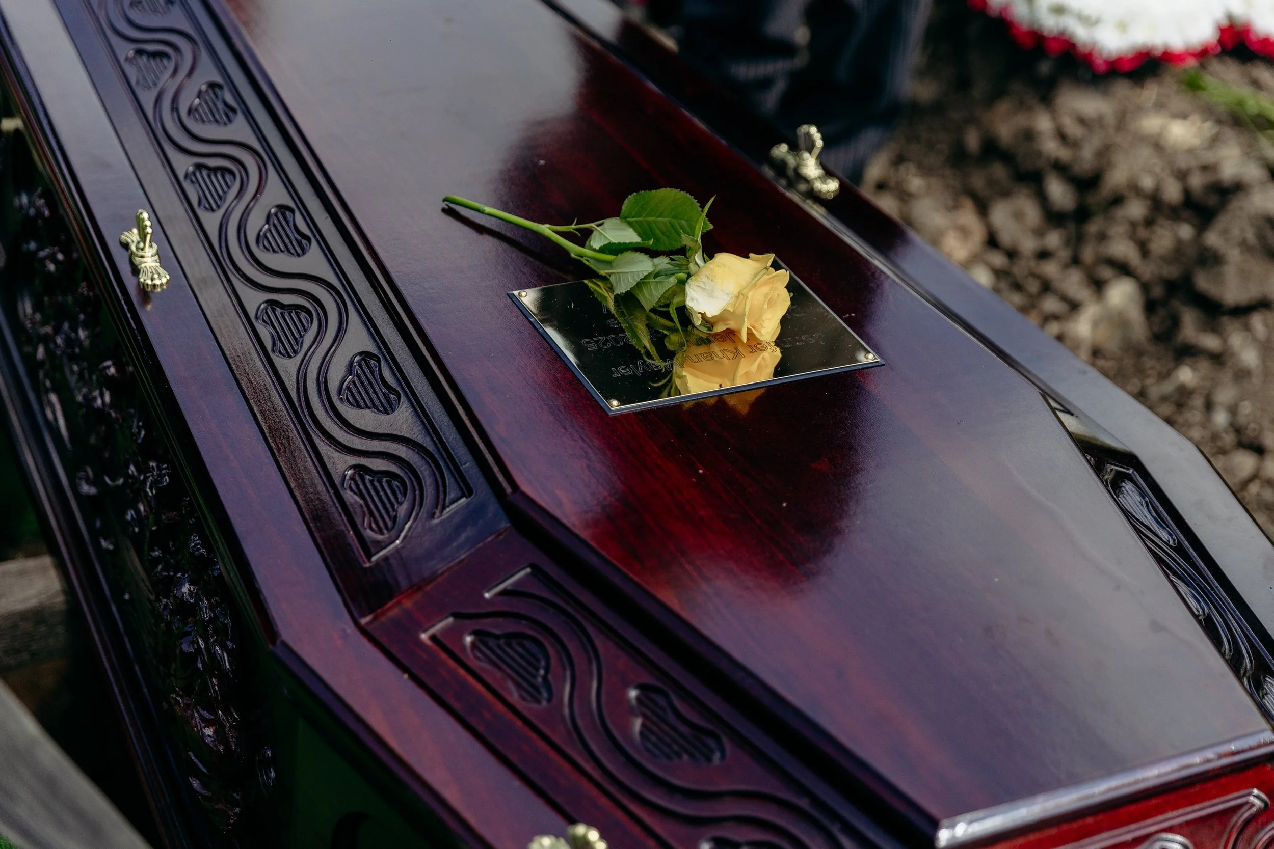 A closed wooden coffin with decorative metal hardware, lying on a platform, with a yellow rose placed on a small reflective metal plaque on top of it, against a background of soil and flowers.