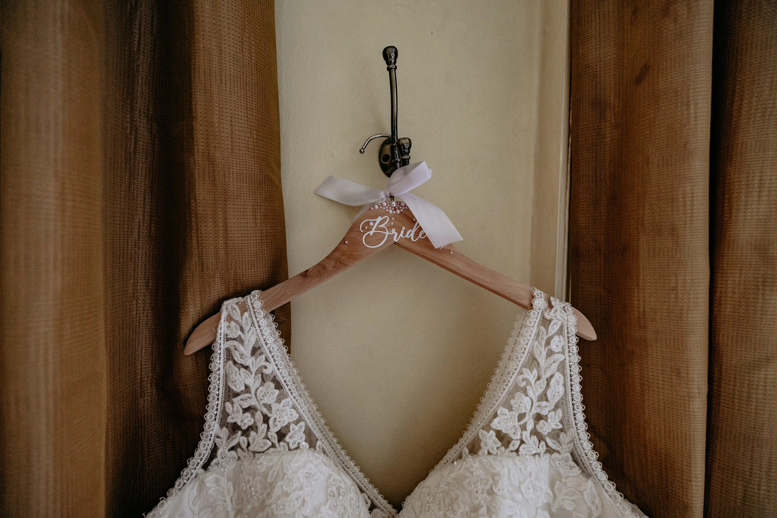 Wedding dress with lace details hanging on a hanger labeled 'Bride' with a white ribbon bow.
