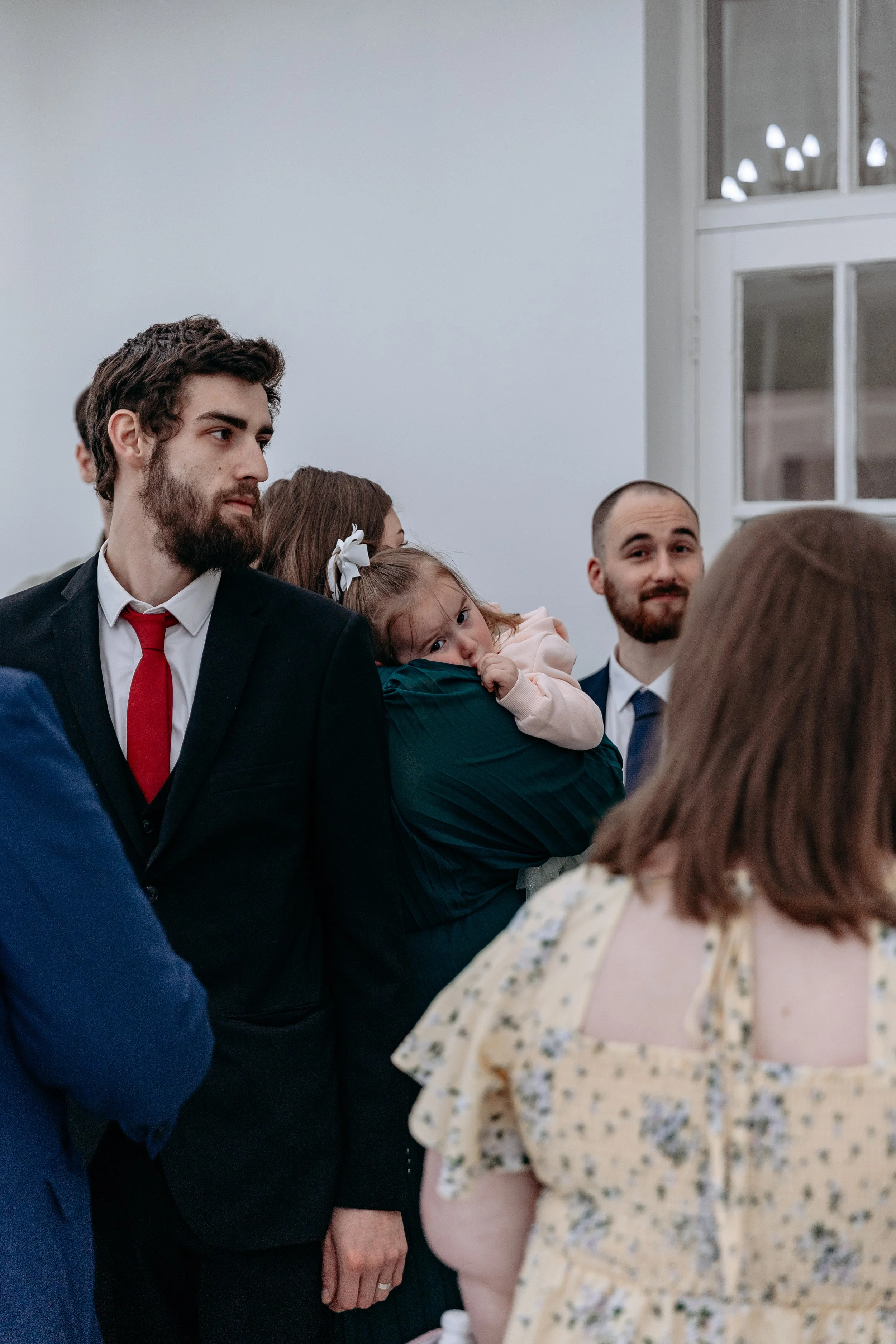 A group of people at a social gathering indoors, with two men dressed in suits, a woman holding a young girl with bows in her hair, and another woman in a floral dress, engaging in conversation or listening attentively.