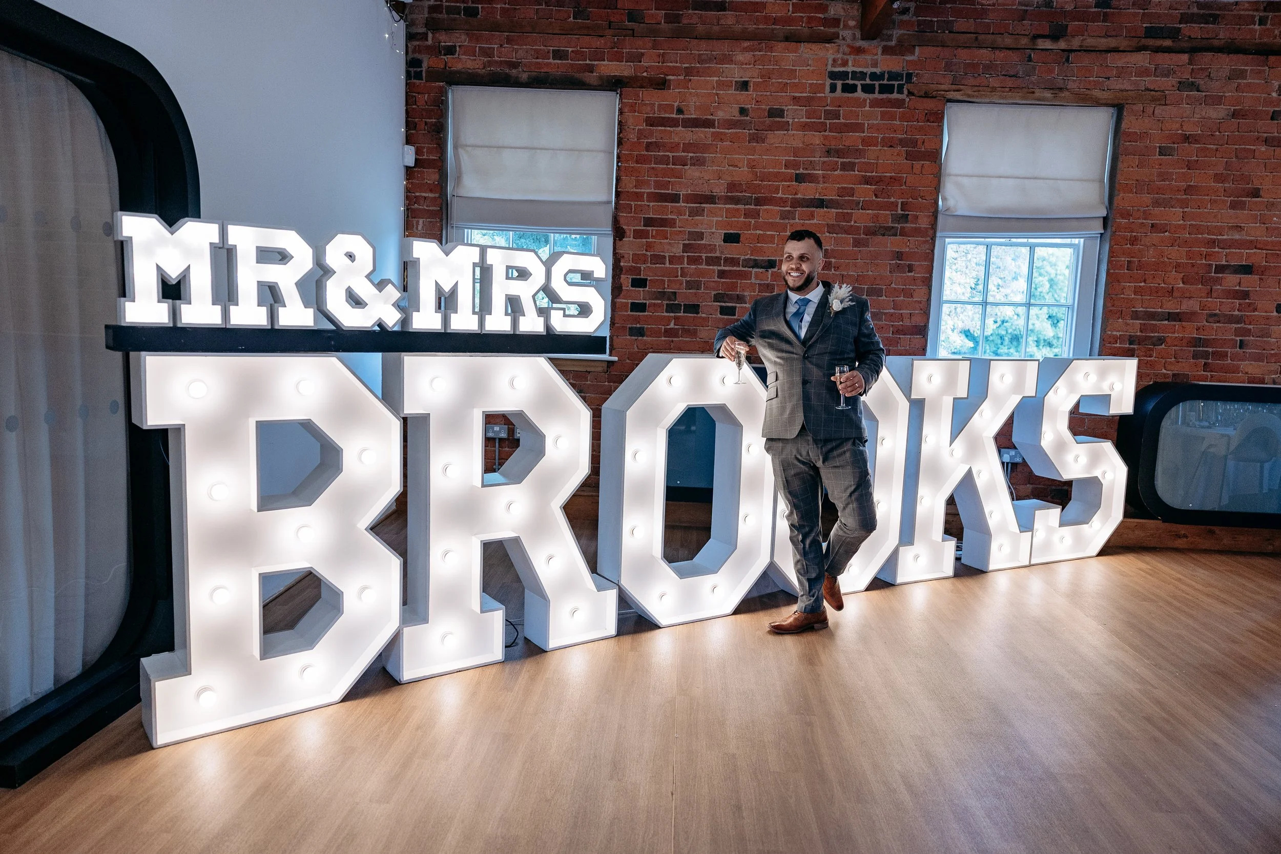 A man in a gray suit dancing with a glass of wine in front of large illuminated letters that spell 'MR & MRS BROOKS' at a wedding reception venue with brick walls and windows.