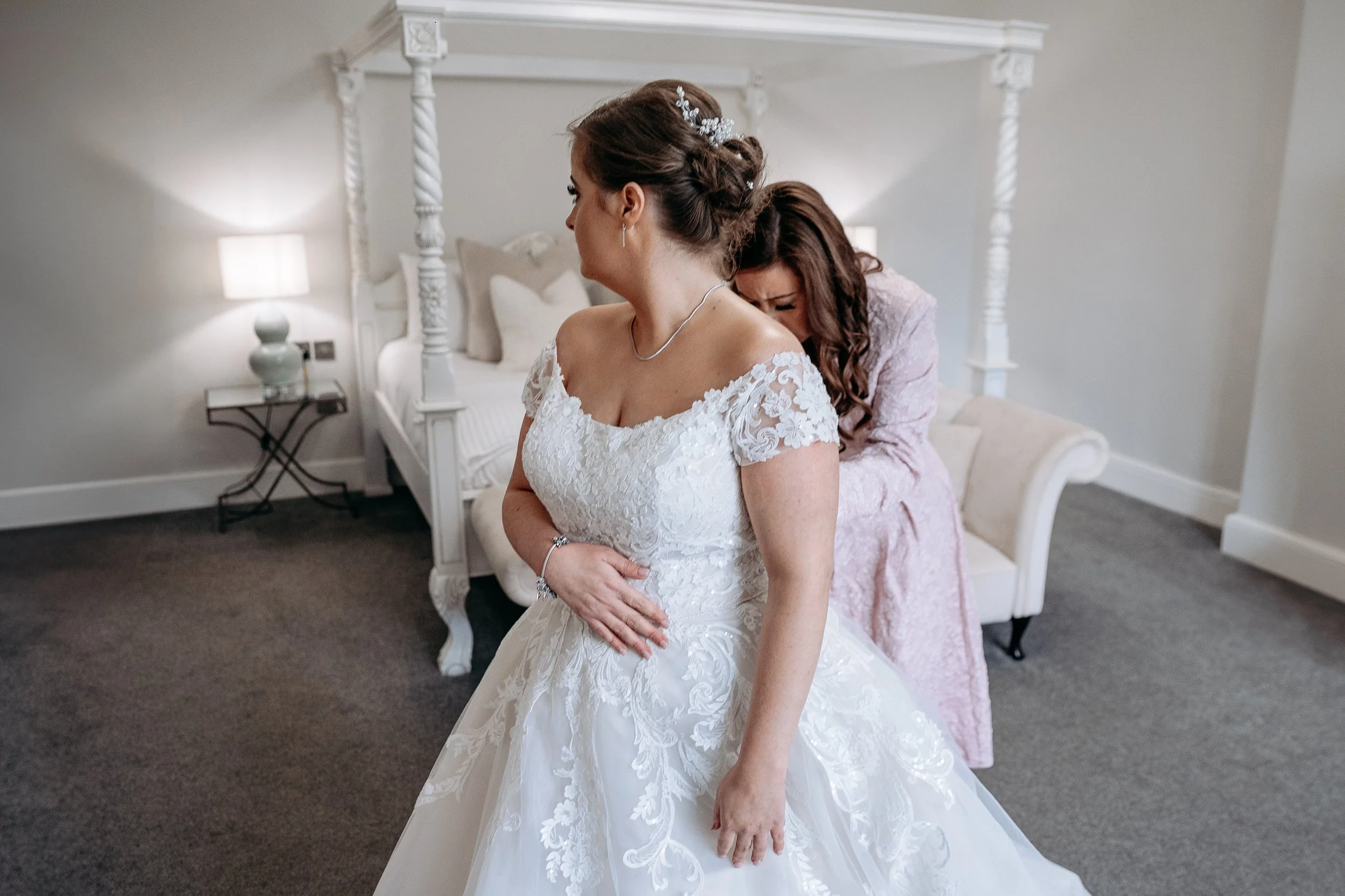 A bride in a white wedding gown with floral lace details stands in a bedroom, while a woman dressed in pink helps her with her dress. The bride has her hand on her stomach, and the woman behind her appears to be adjusting or fixing the gown.