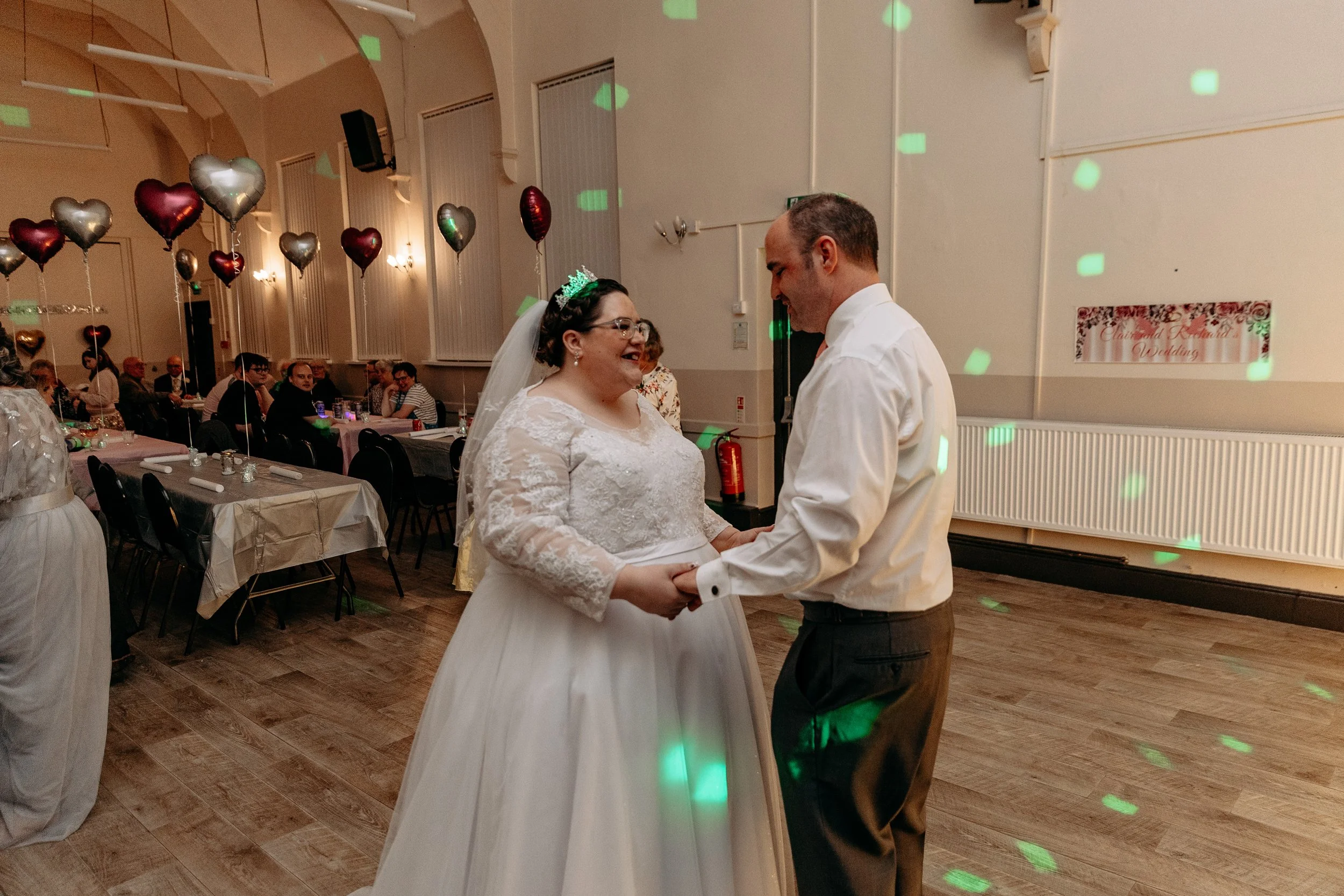 A bride and groom dance together in a reception hall decorated with heart-shaped balloons and a sign that reads "Grand Rea..." in the background. Guests are seated at tables, with some watching the couple dance.