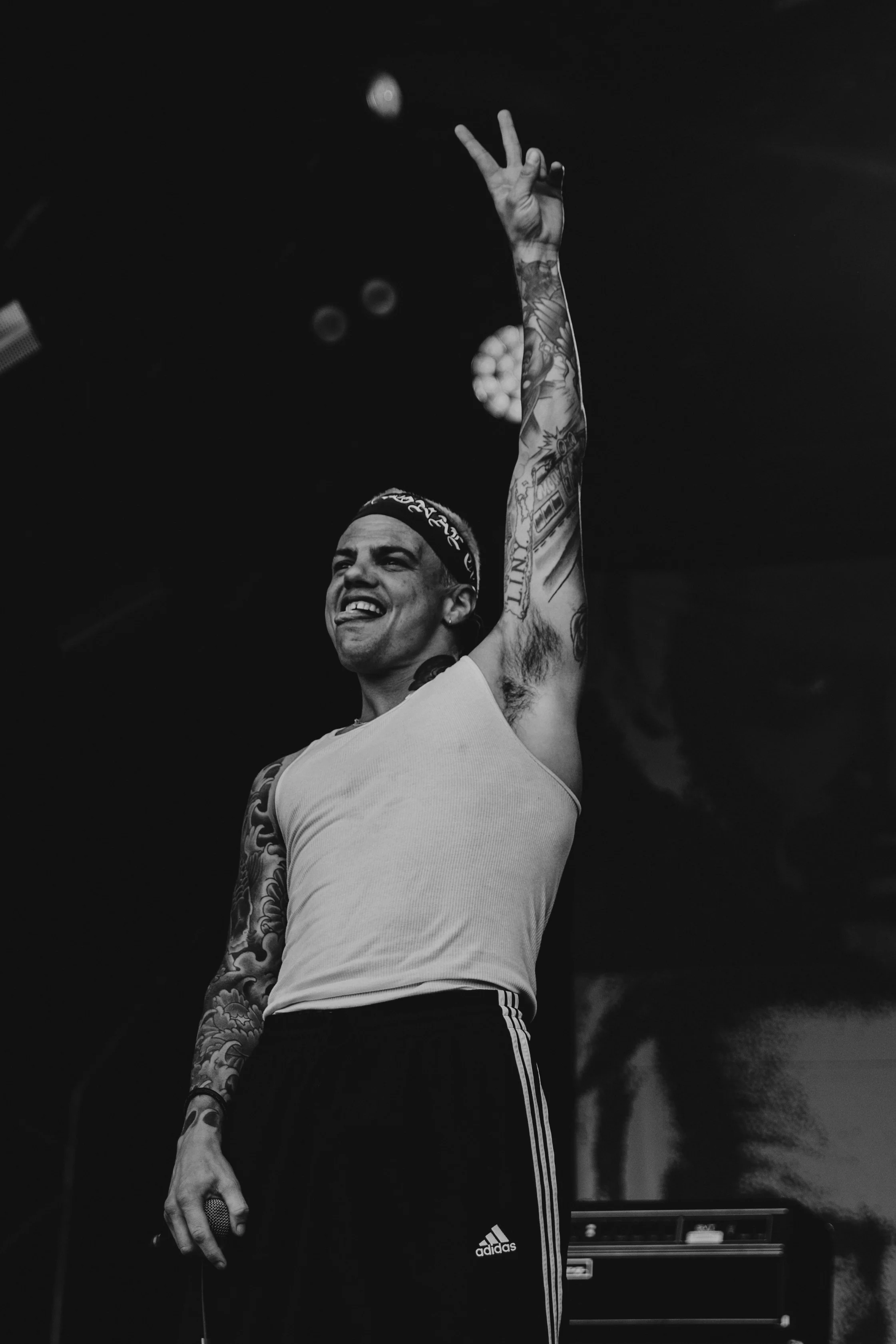 A man with tattoos on his arms, wearing a headband, white sleeveless shirt, and Adidas sweatpants, is holding a microphone in one hand and raising the other with a peace sign gesture during a performance or concert.