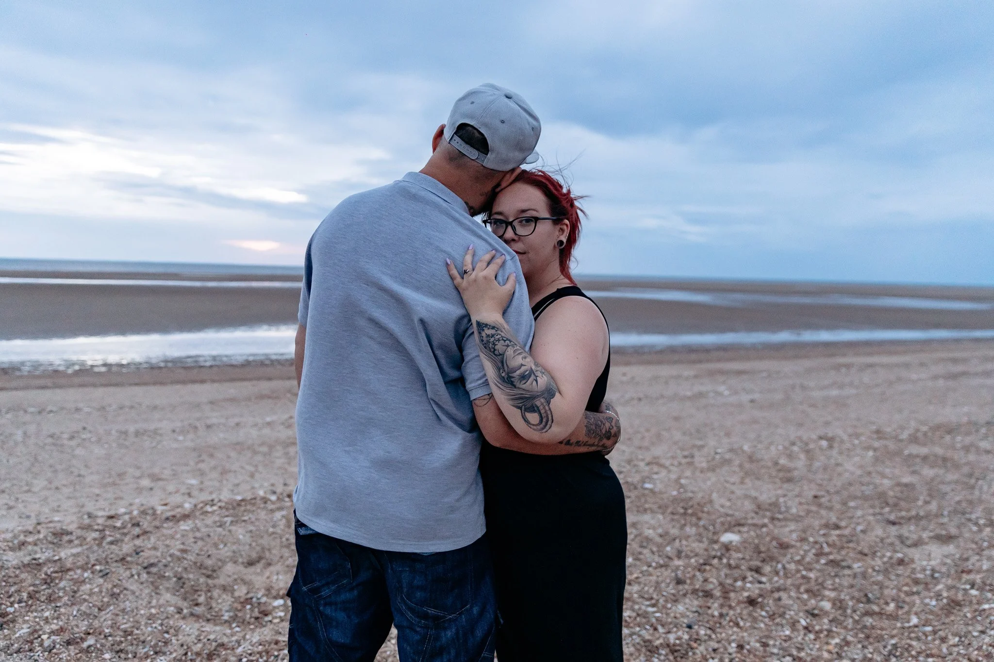 A couple embracing on a sandy beach during dusk, with the man wearing a light gray cap and the woman with red hair and glasses.