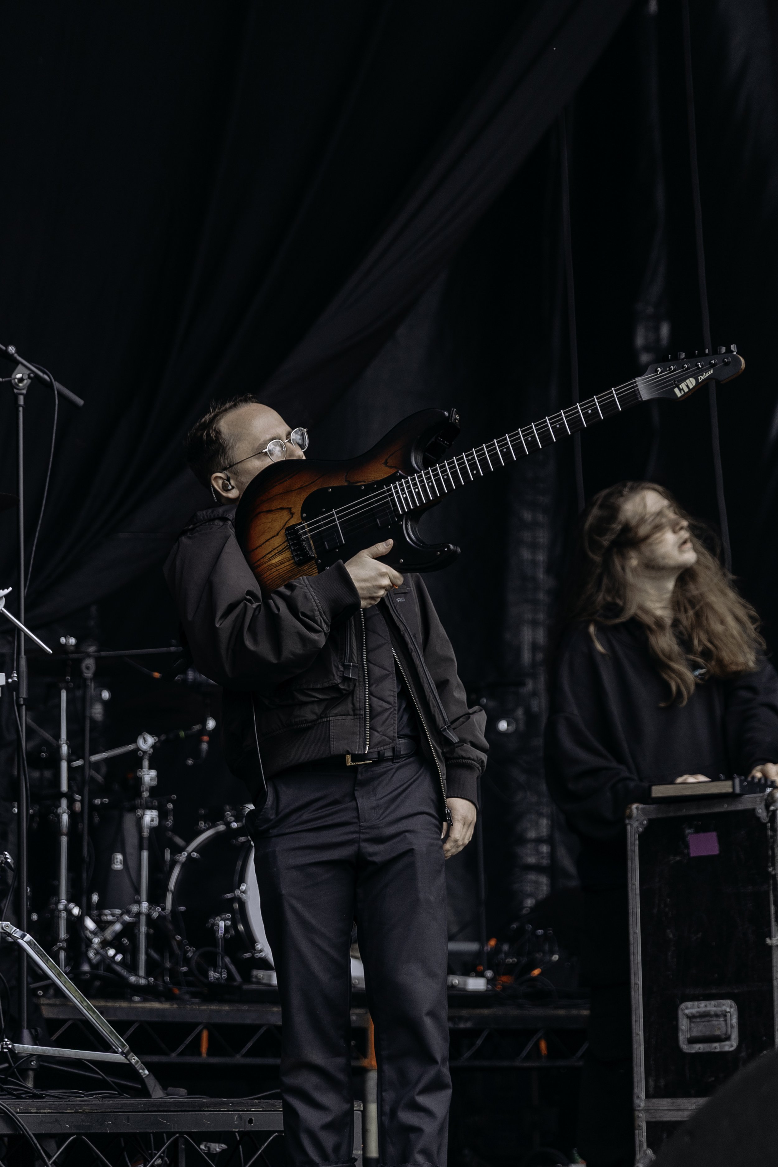Man playing an electric guitar on stage with a dark curtain backdrop and drum set in the background.