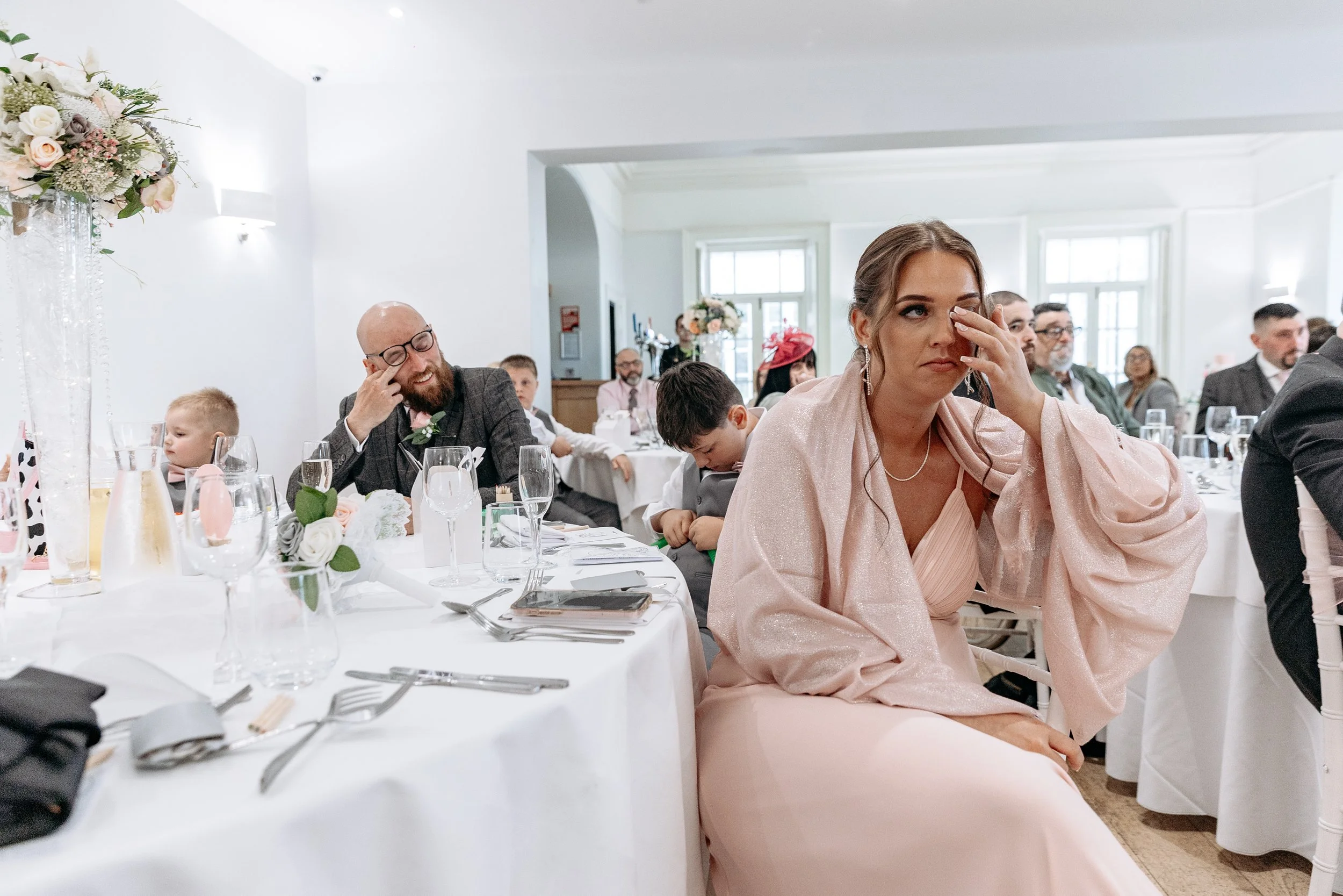 A woman in a pink dress is sitting at a banquet table, wiping away tears, while a group of people, including children, are seated behind her, some laughing and others appearing emotional, in a well-lit event hall with white walls and floral decoratio