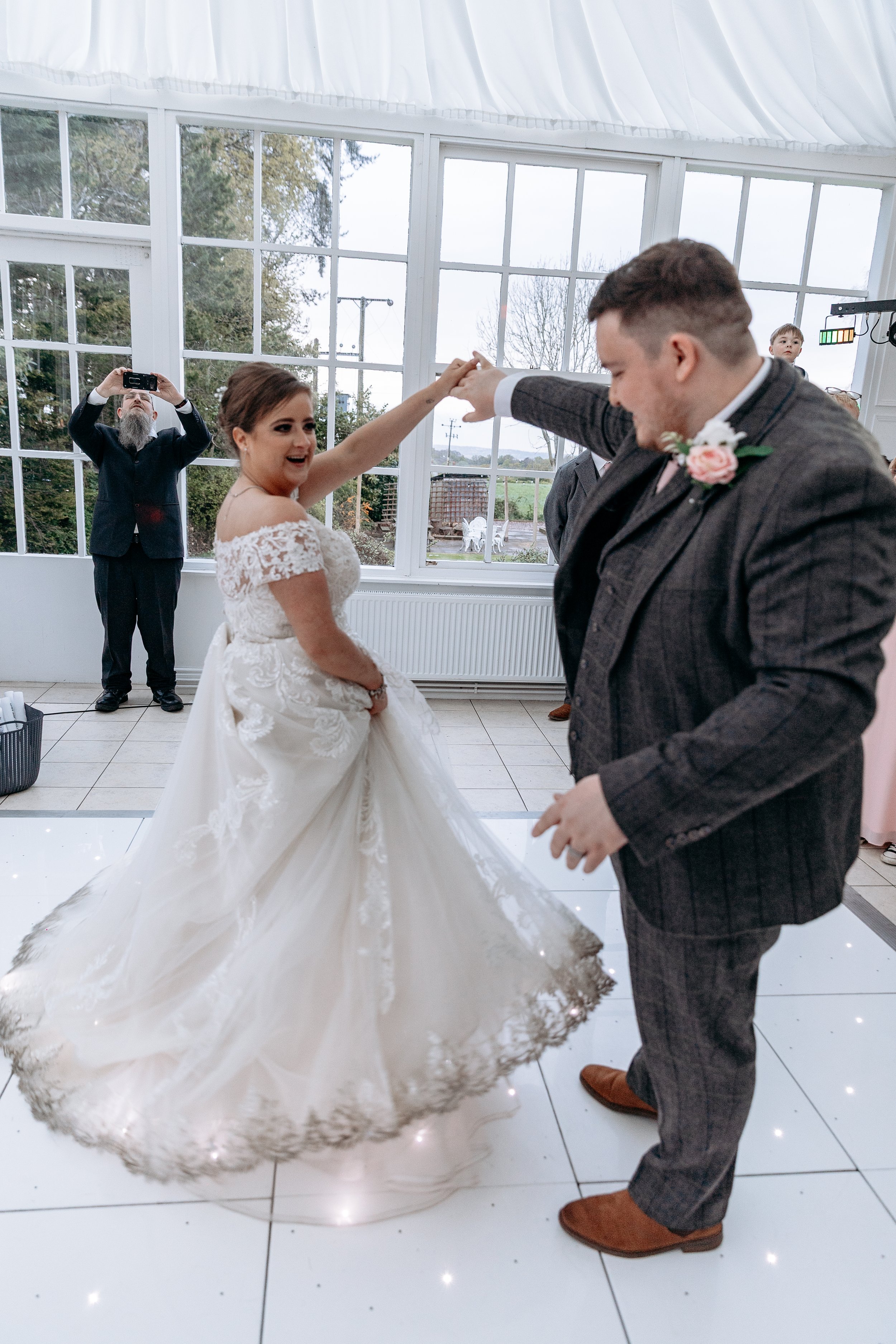 Bride and groom dancing at their wedding reception, with guests taking photos indoors.