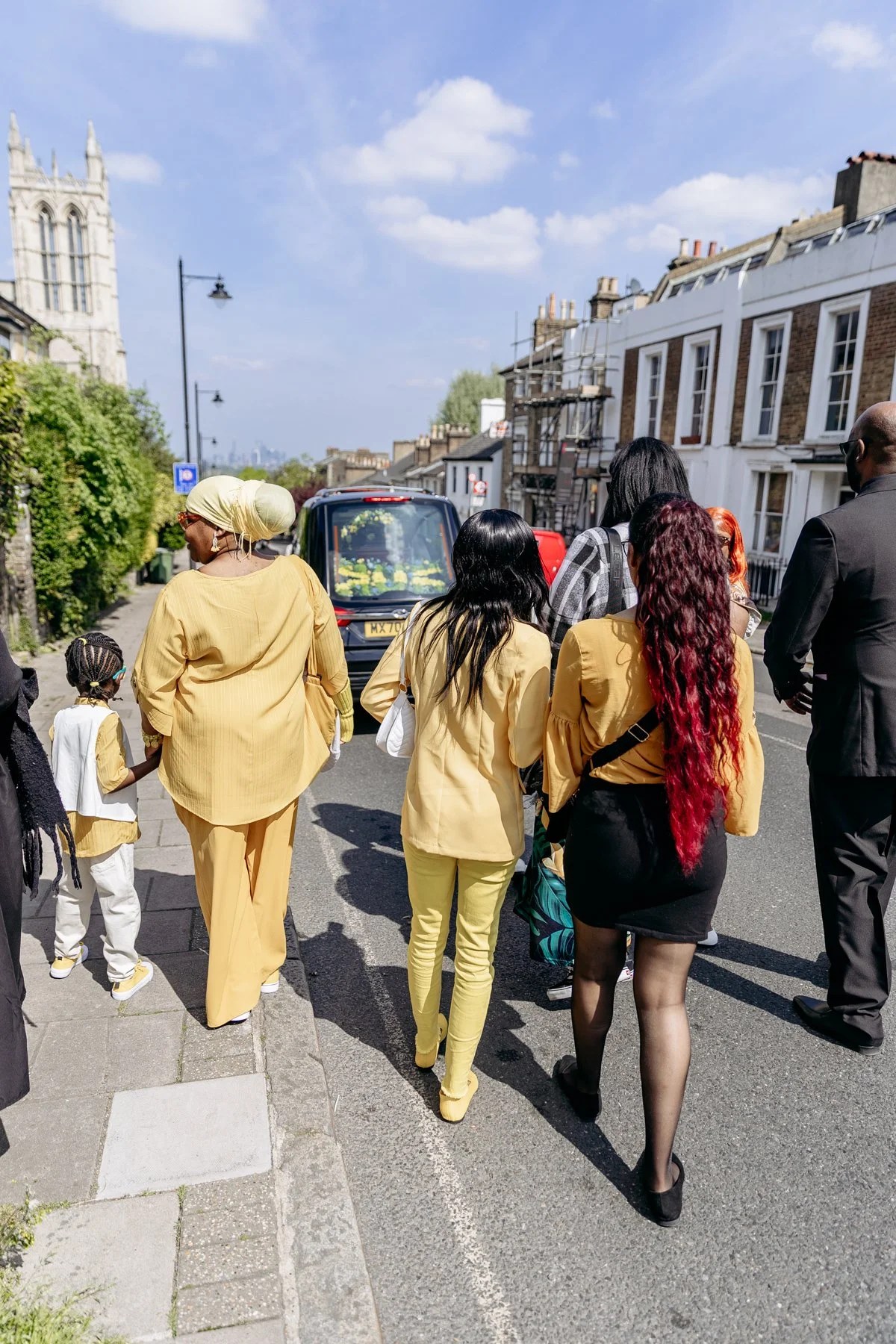 Group of diverse people walking on a street, some wearing yellow clothing, with a black vehicle and old buildings in the background on a sunny day.