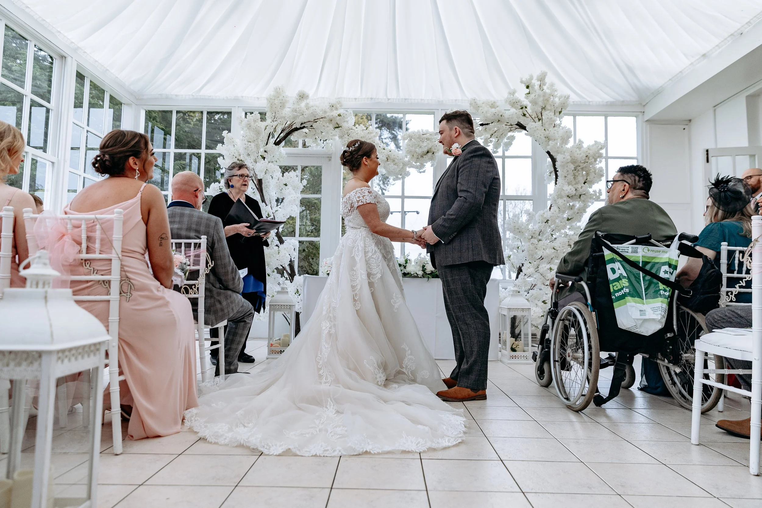 A bride and groom holding hands during their wedding ceremony inside a bright, glass-enclosed venue with white floral decorations and seated guests.