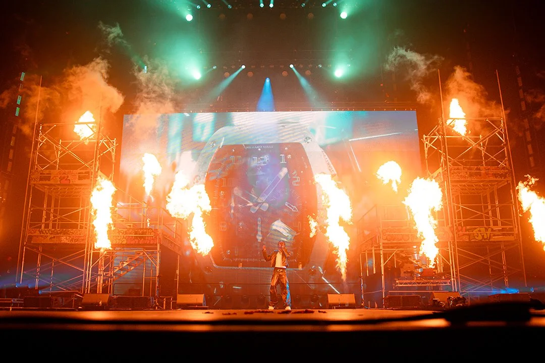 Concert stage with pyrotechnics, large screen in background displaying a watch.