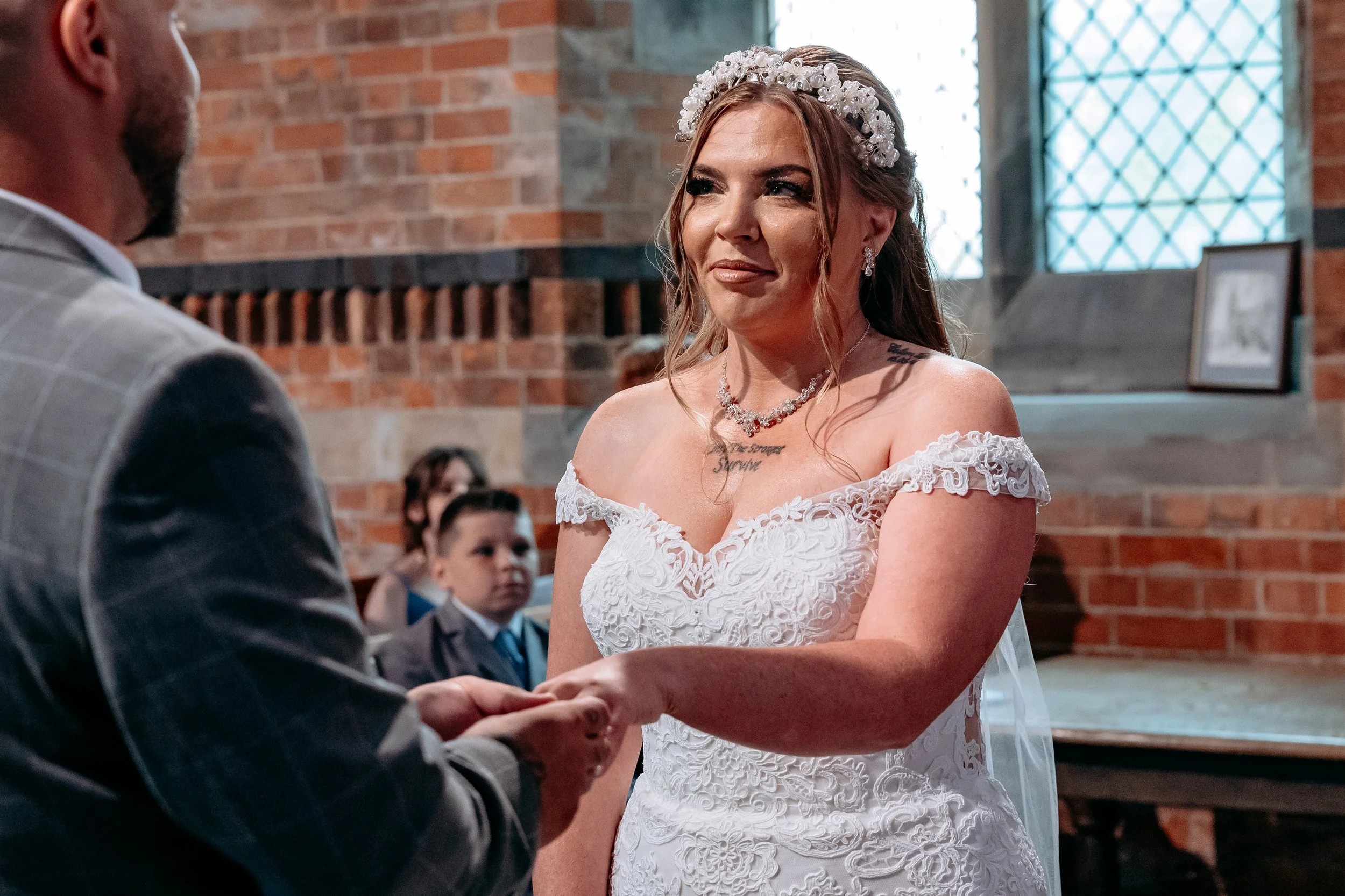A bride in a white lace wedding dress holding hands with a groom during a wedding ceremony inside a brick building.
