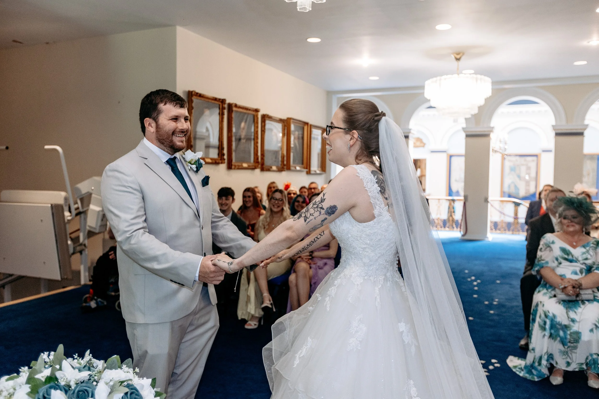 A bride and groom holding hands and smiling during their wedding ceremony in a decorated indoor venue with guests seated and watching.