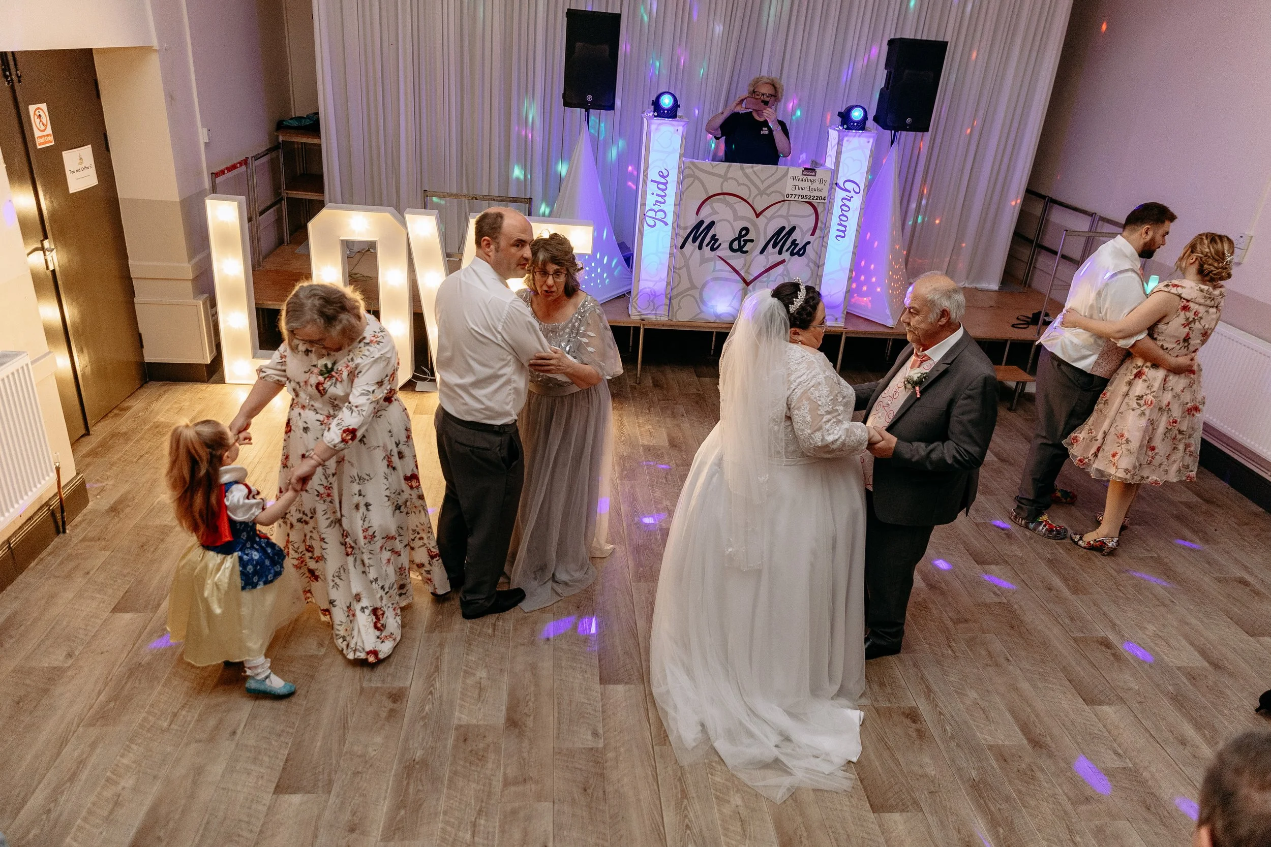 Guests dancing at a wedding reception with a bride and groom in the center, a DJ booth with lights in the background, and large illuminated letters spelling out 'Love' nearby.